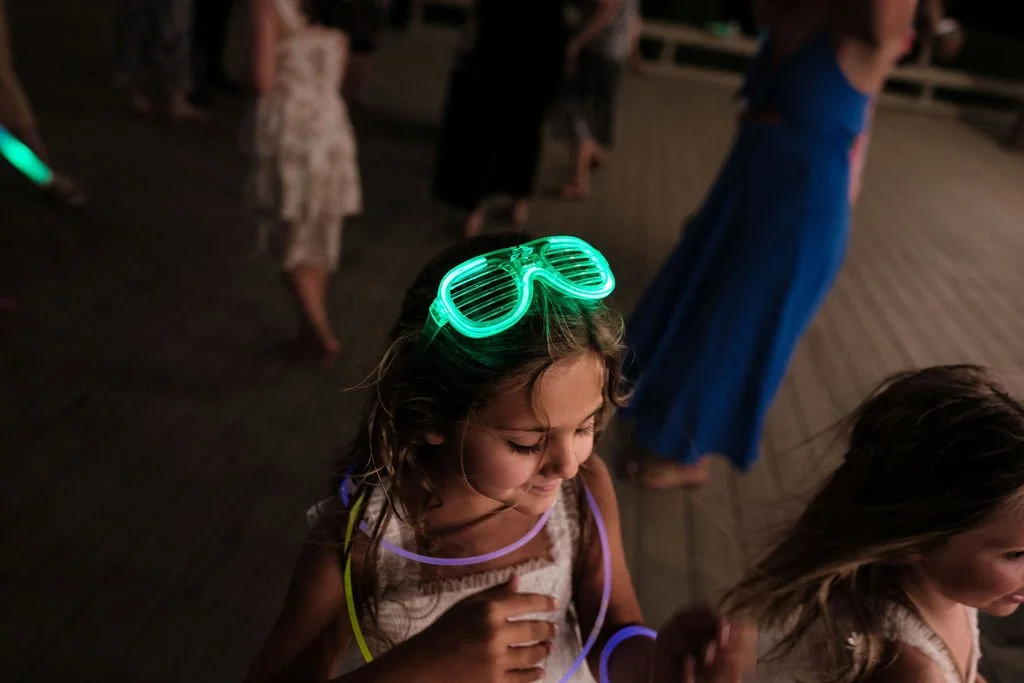 A young girl wearing glowing green shutter shades and glow-in-the-dark necklaces at a nighttime outdoor event.