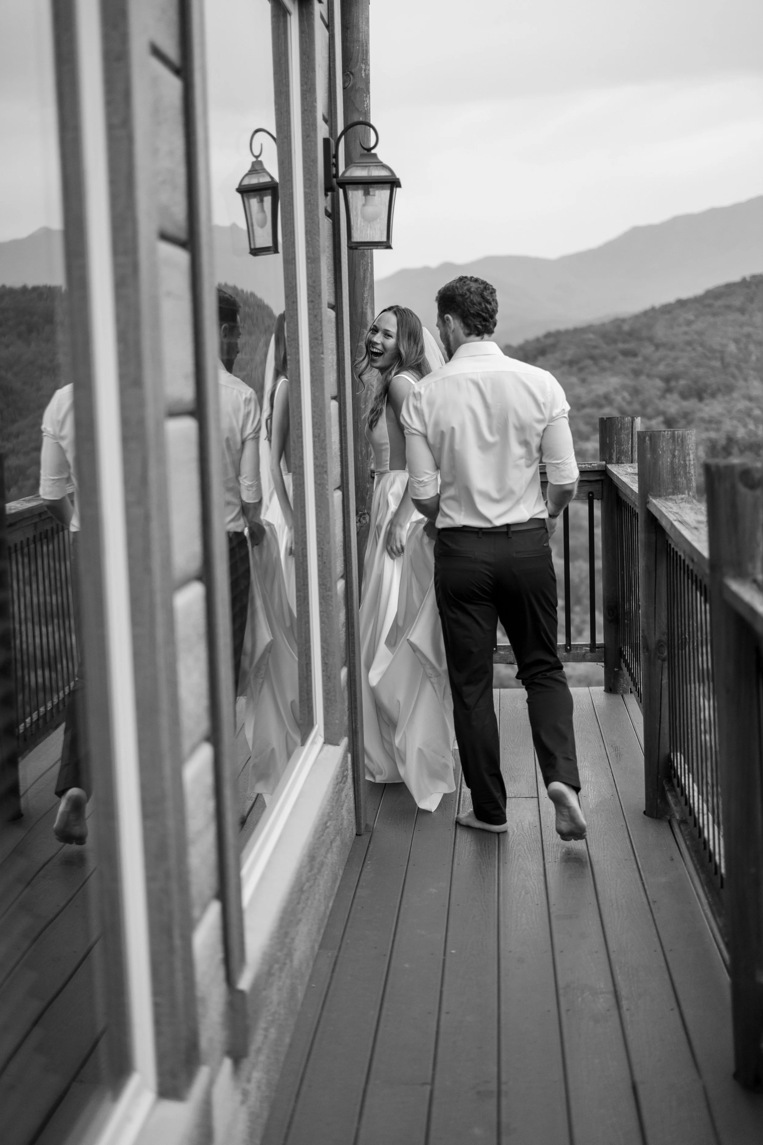 A black-and-white photo of a couple on a balcony with mountains in the background. The woman is in a wedding dress and is laughing, while the man is in a dress shirt with rolled-up sleeves, standing barefoot with his back to the camera.