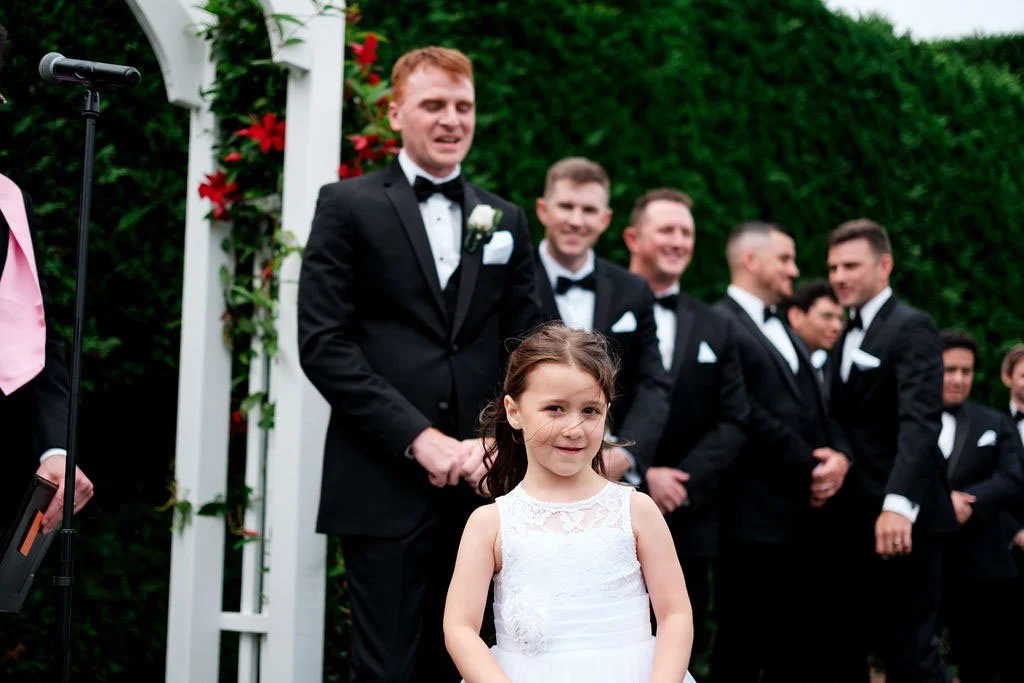 A wedding ceremony outdoors, featuring a young girl in a white dress standing in front of a group of men in tuxedos, with a floral arch behind them.