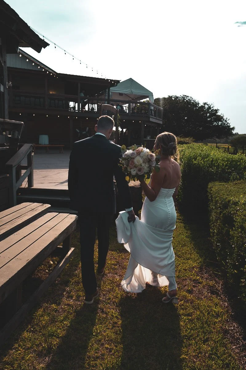 A bride in a white wedding gown holding a bouquet of flowers, walking hand-in-hand with a groom in a black suit outside during sunset.
