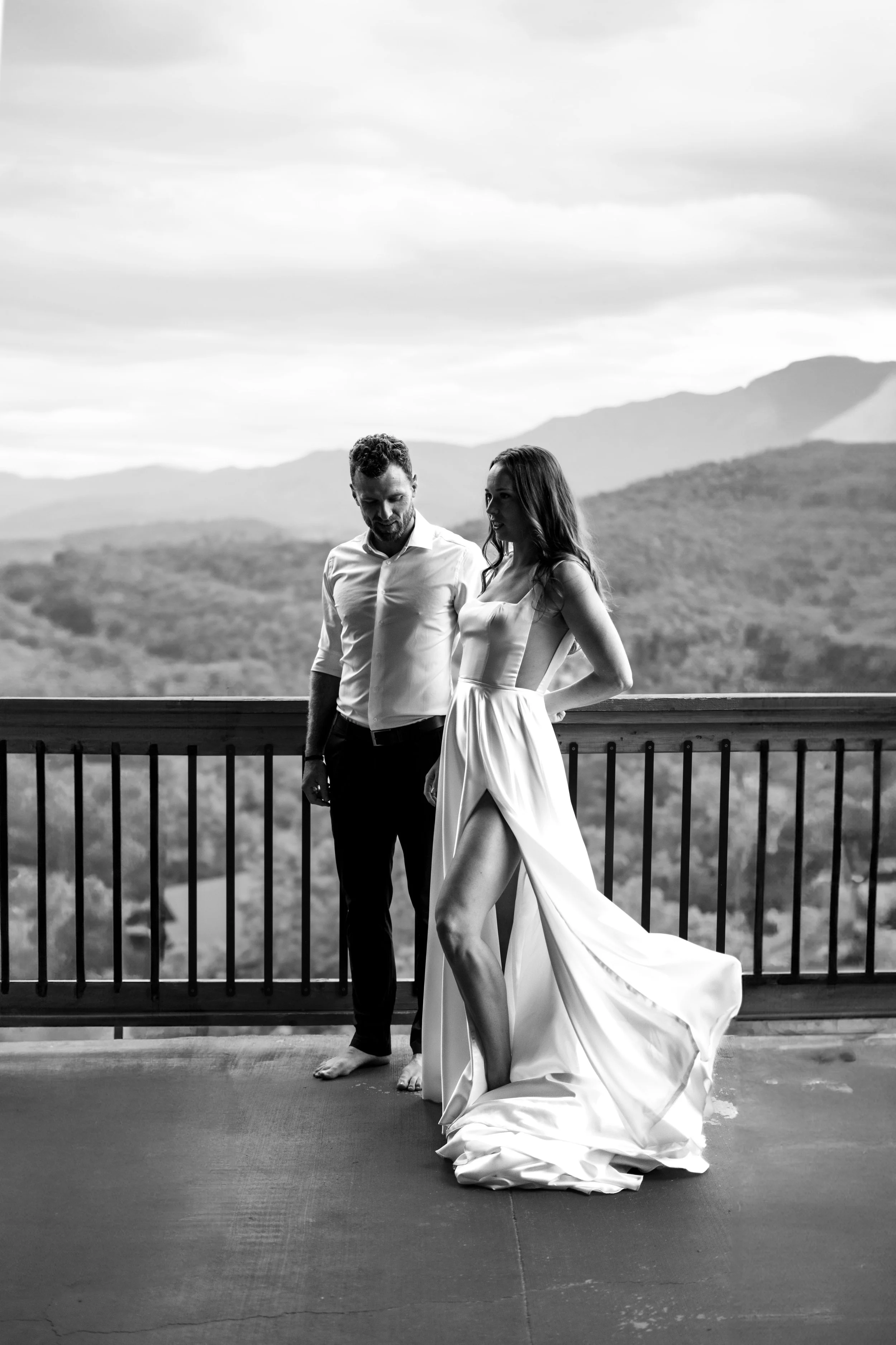 A black and white photo of a man and woman on a balcony with a mountain landscape in the background. The woman is dressed in a flowing wedding gown with a high slit, and the man is in a white shirt and dark pants. They are barefoot and looking at eac