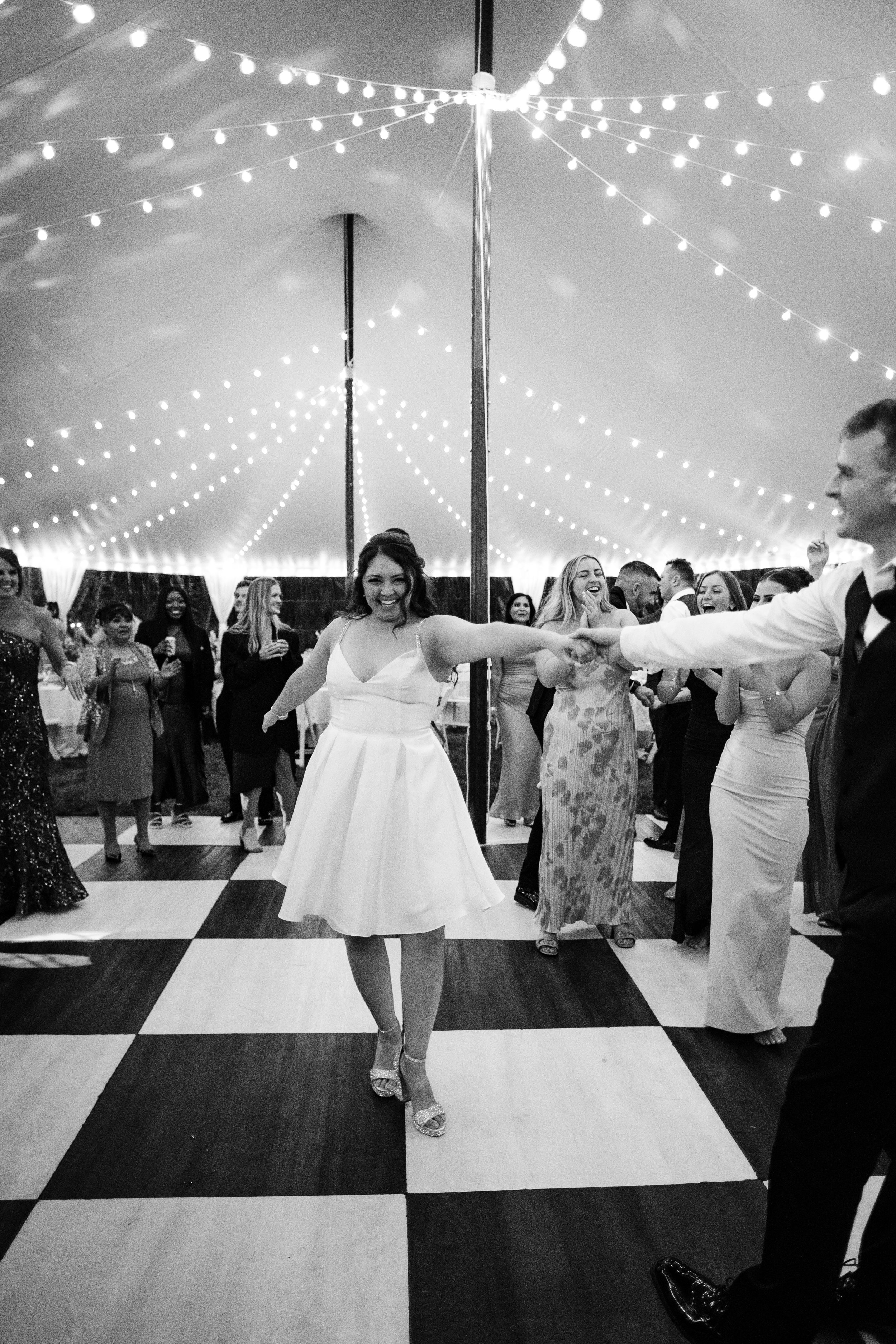 Black and white photo of a woman in a white dress dancing and holding hands with a man in a suit at a wedding reception under a tent decorated with string lights, with several guests watching and enjoying the celebration.
