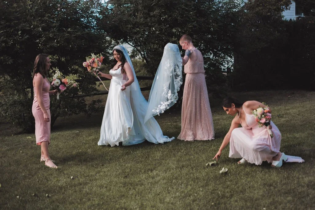 A bride in a white wedding dress holding a bouquet of flowers stands on a grassy lawn, surrounded by four women, one of whom is bending down to pick up flowers, while others hold bouquets, with trees in the background.