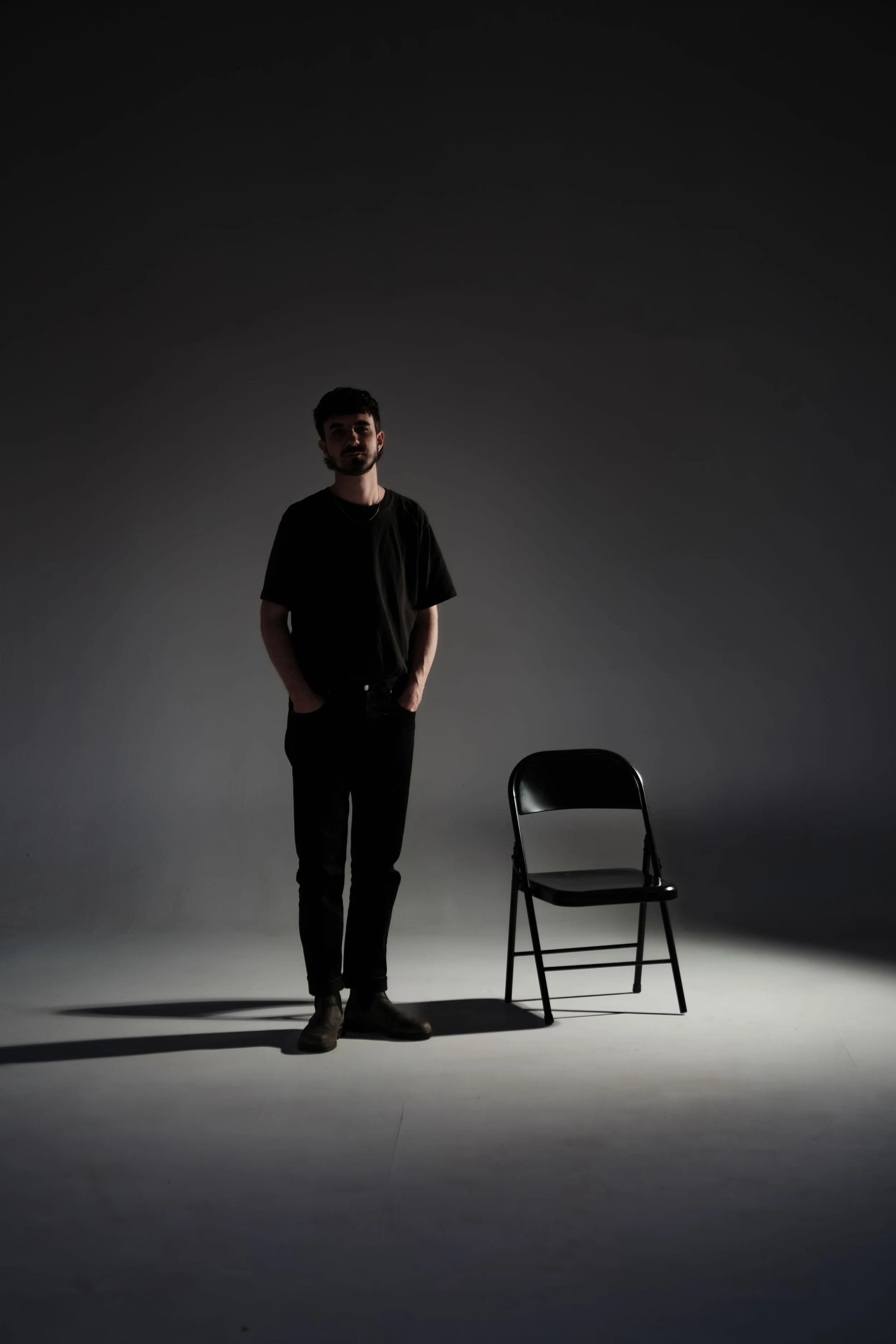 A young man standing with hands in pockets next to a black chair on a dimly lit studio background.