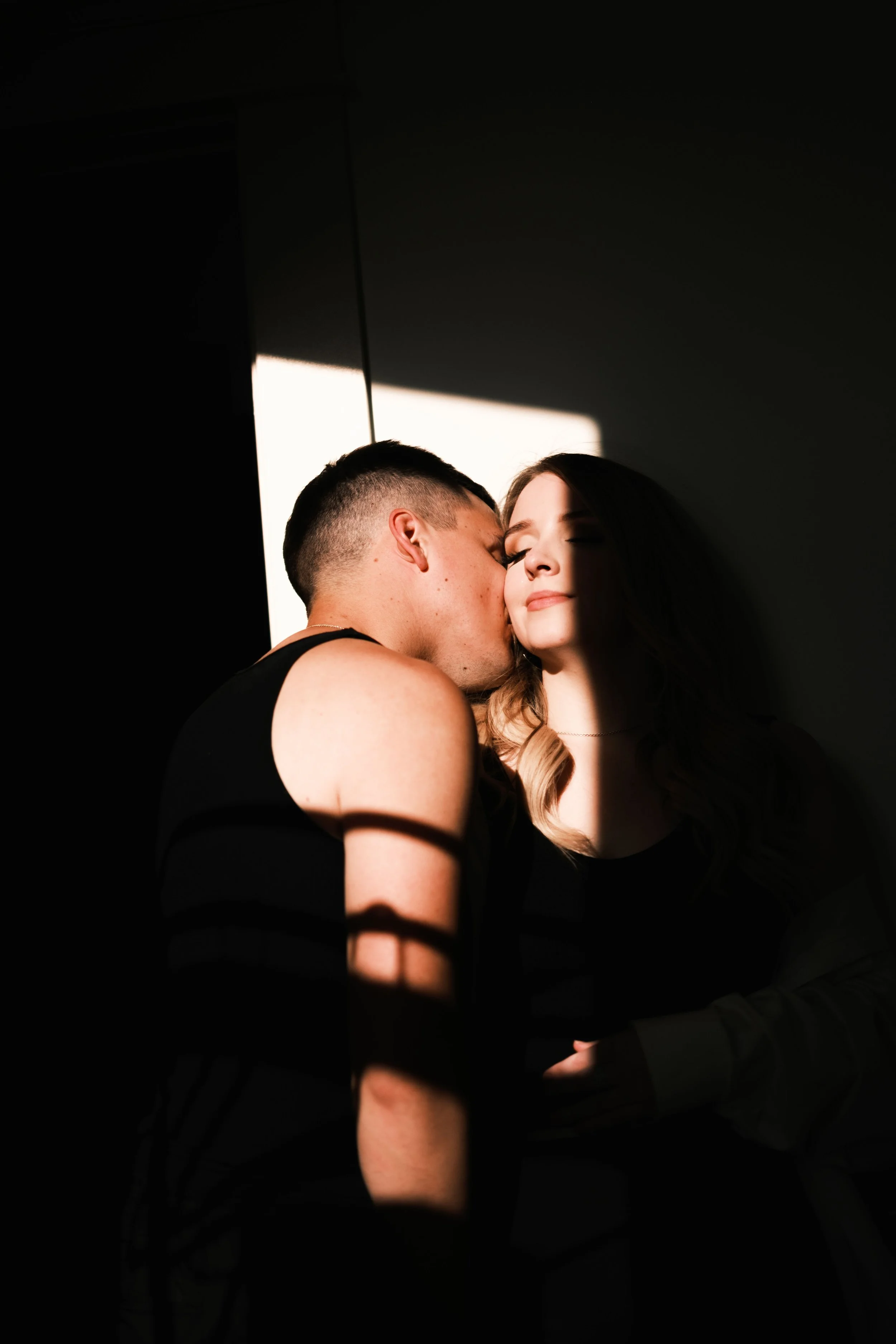 A man kissing a woman on the cheek in a dark room with sunlight streaming through a nearby window.