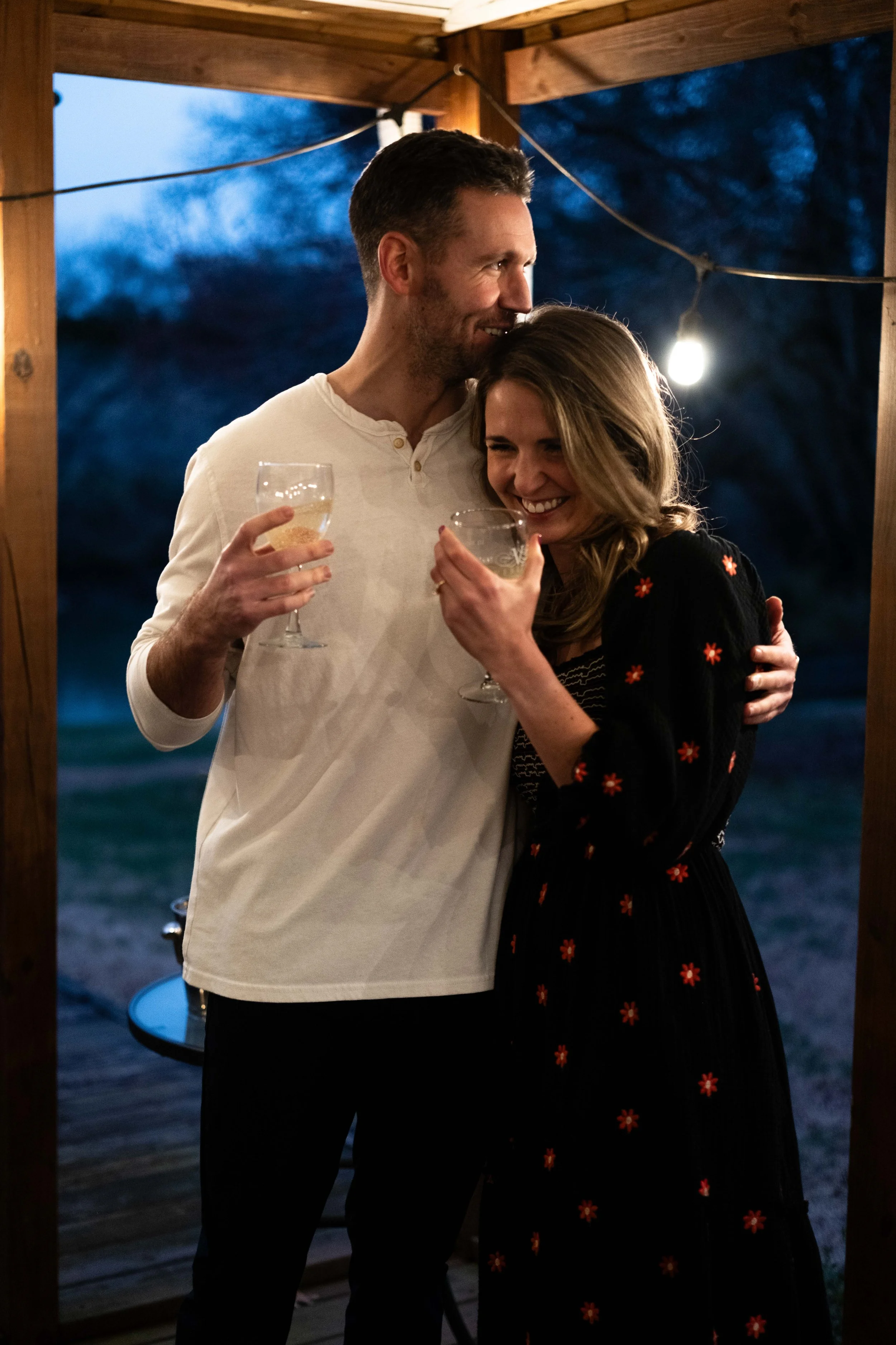A man and woman sharing a happy moment, hugging and holding drinks, under a wooden roof with string lights at dusk.