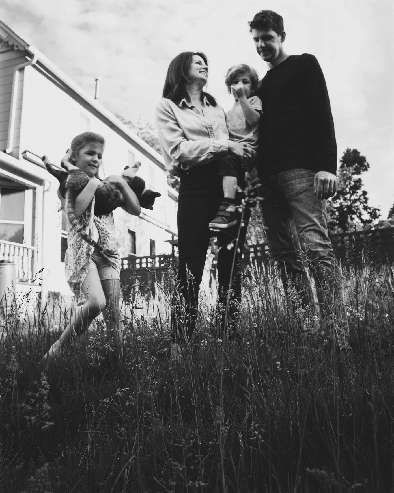 A family of four with two young girls and two adults, standing outside in a grassy yard near a house, smiling and enjoying each other's company on a partly cloudy day.