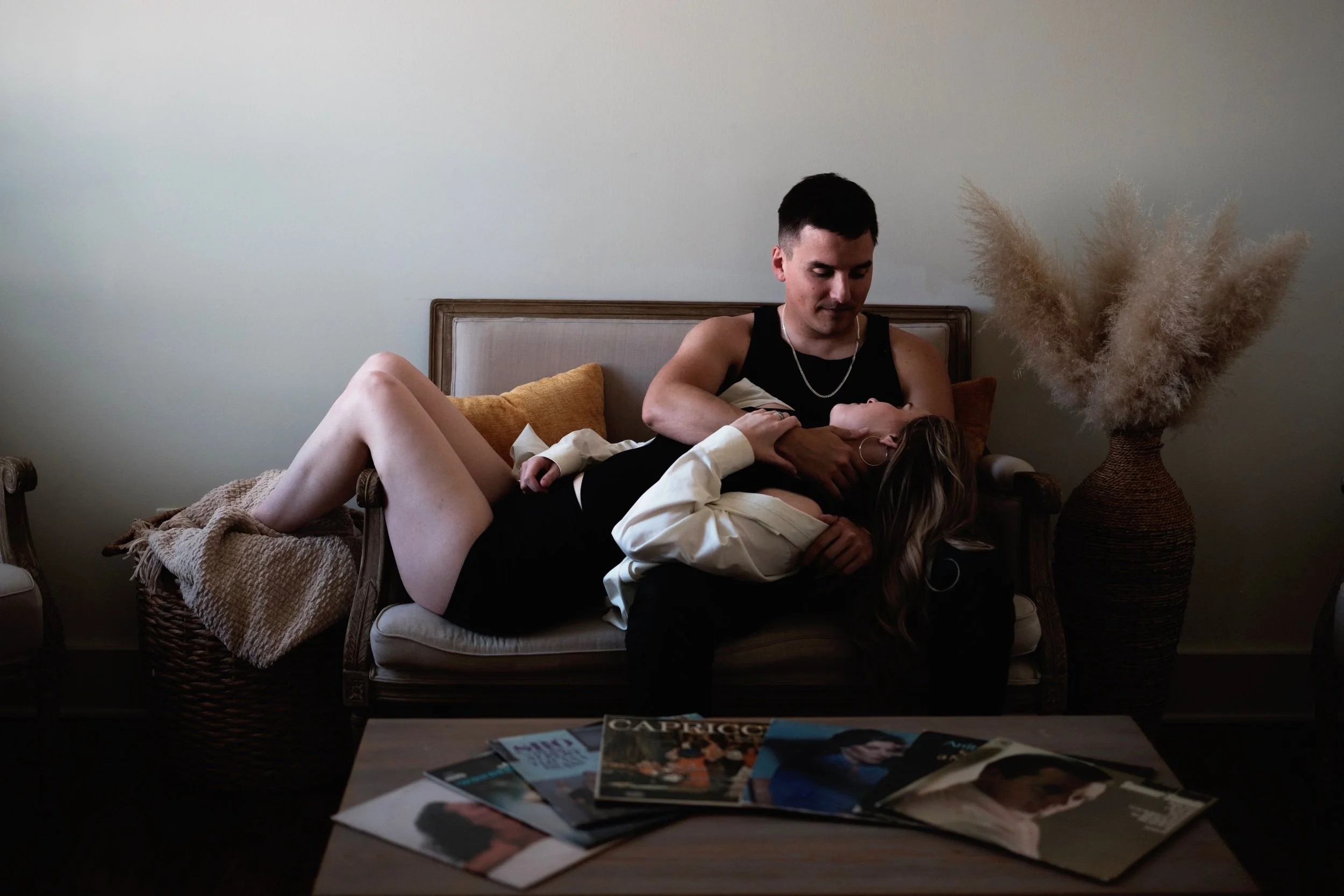 A man and a woman lie on a vintage sofa. The man, wearing a black sleeveless shirt, is sitting up while the woman, dressed in black with a white jacket, is reclining with her head on his lap. There are magazines on a nearby table, and a large vase wi