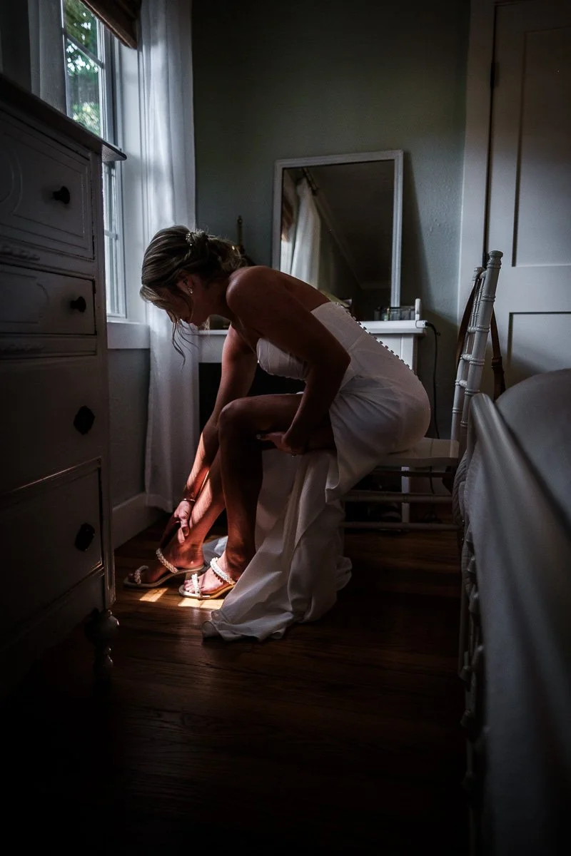 A woman in a white dress sitting on a chair while putting on a sandal near a window in a bedroom.