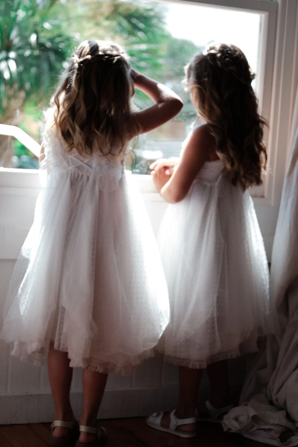 Two young girls in white dresses standing by a window, looking outside.