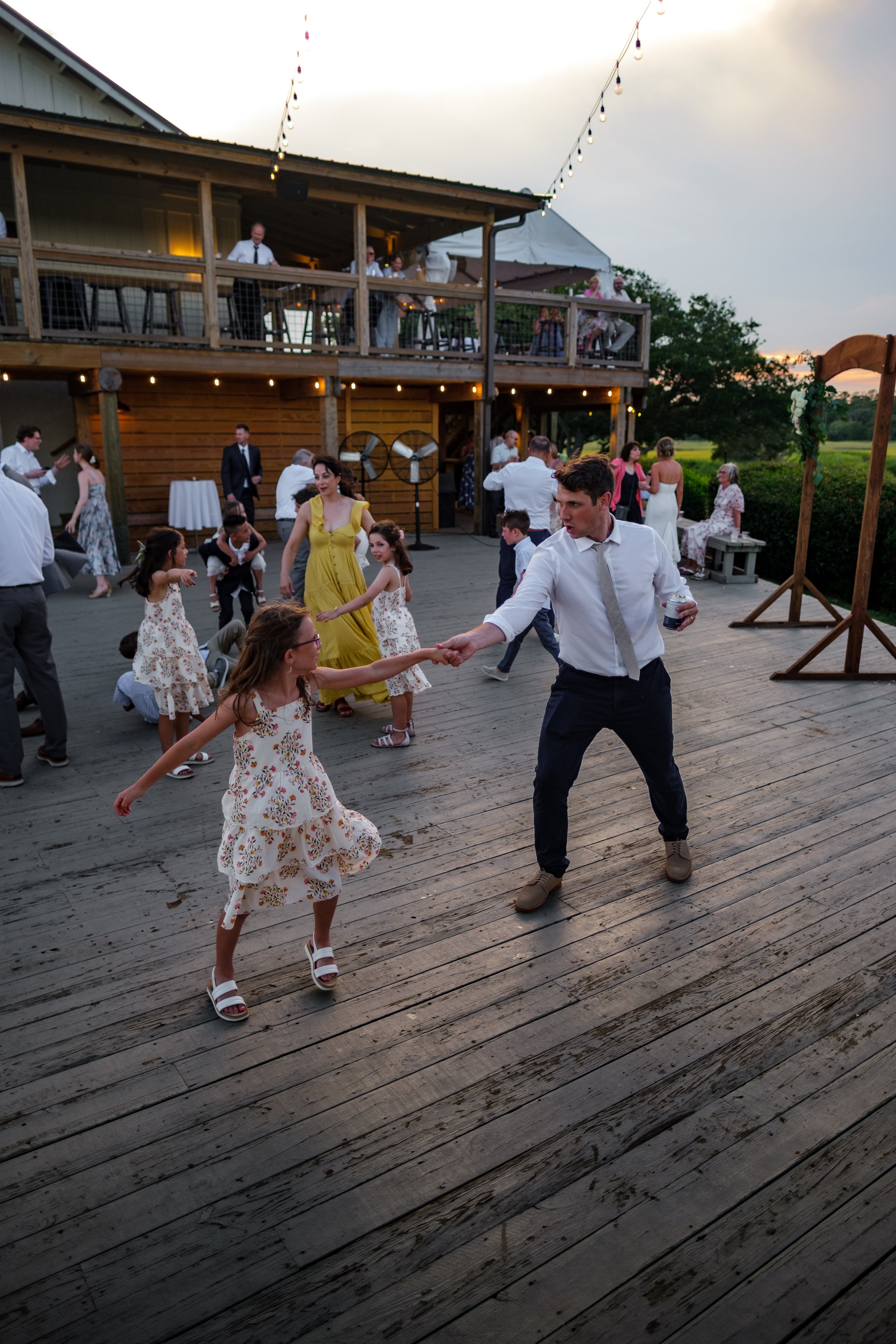 People dancing and socializing on an outdoor wooden deck at a party or celebration during sunset, with a two-story wooden building and string lights in the background.