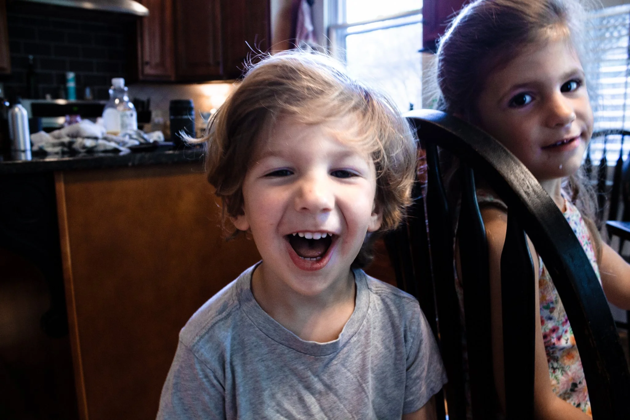 Two children sitting at a dining table, one boy with curly blond hair laughing and a girl with long brown hair smiling, in a kitchen with dark wooden cabinets and a window in the background.