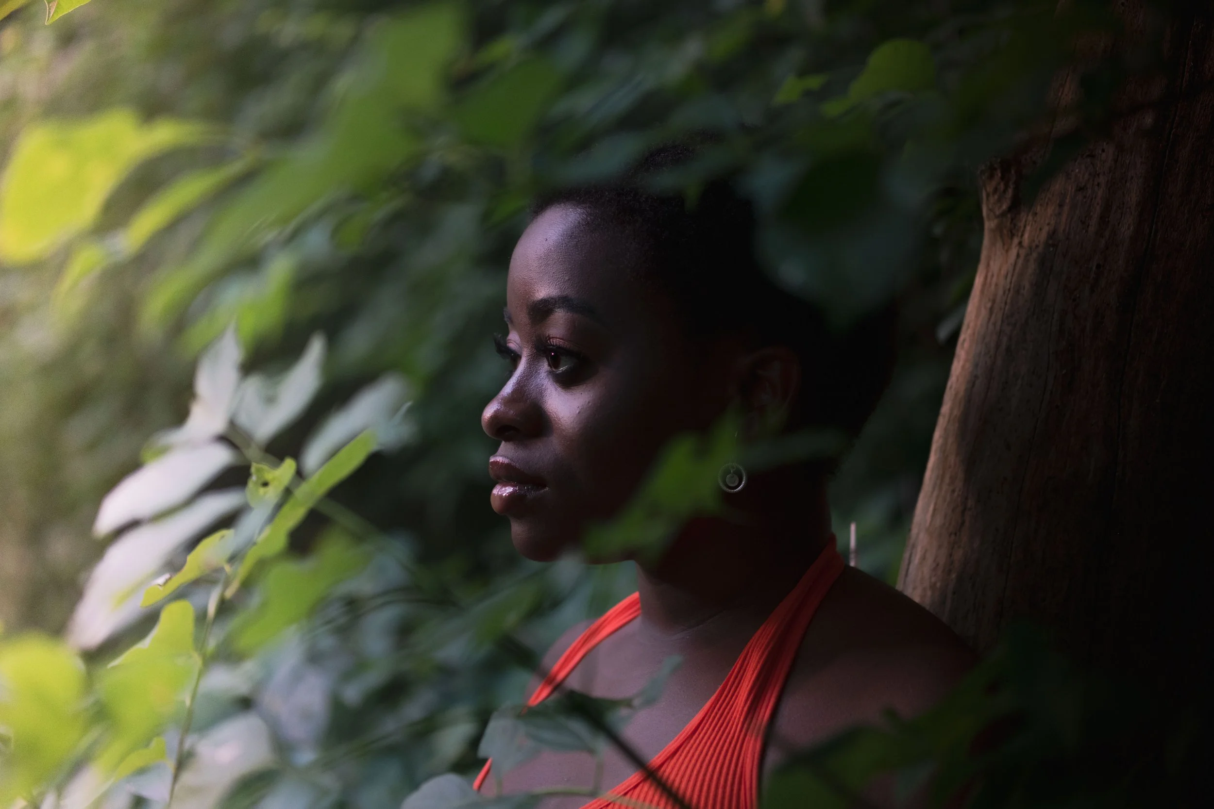 A woman with dark skin and detailed makeup, wearing an orange top, leaning against a tree in a lush green forest, surrounded by leaves.