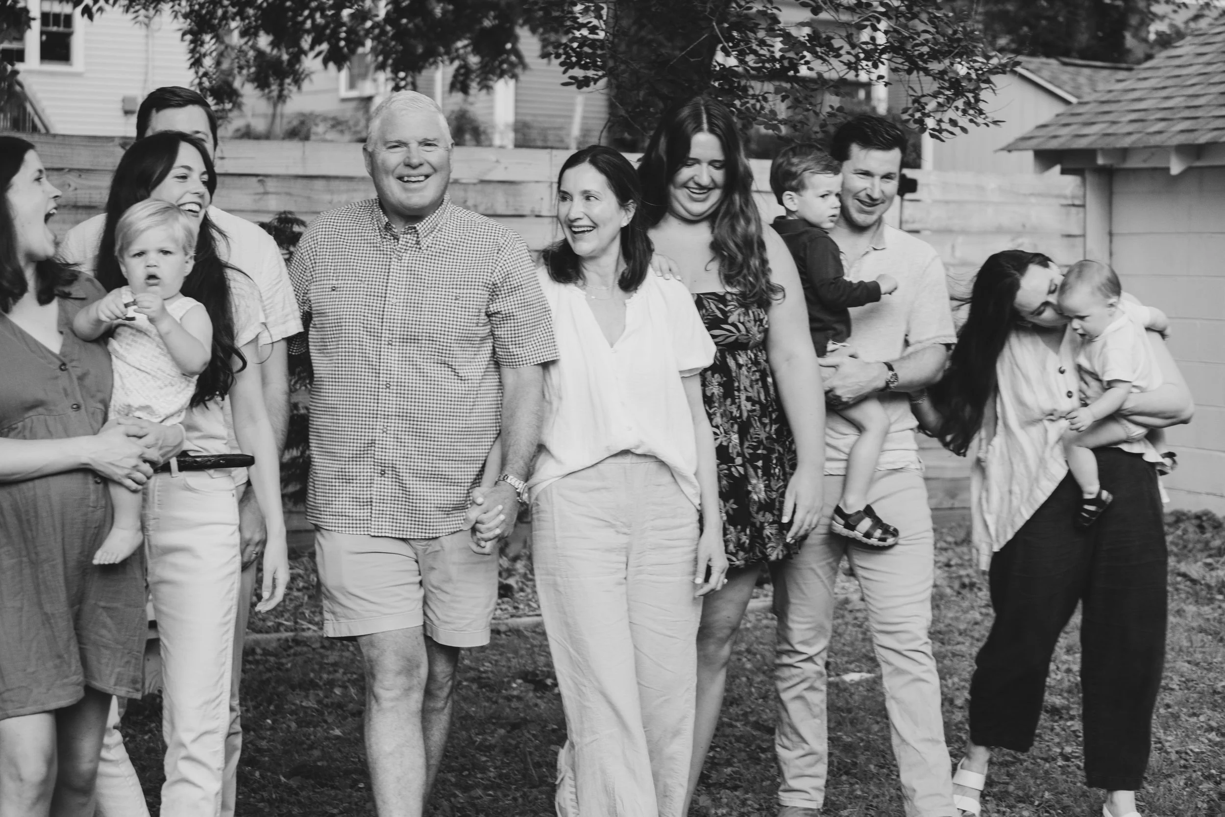 A black and white photo of a smiling multi-generational family gathering outdoors, with people holding children and standing in a row.