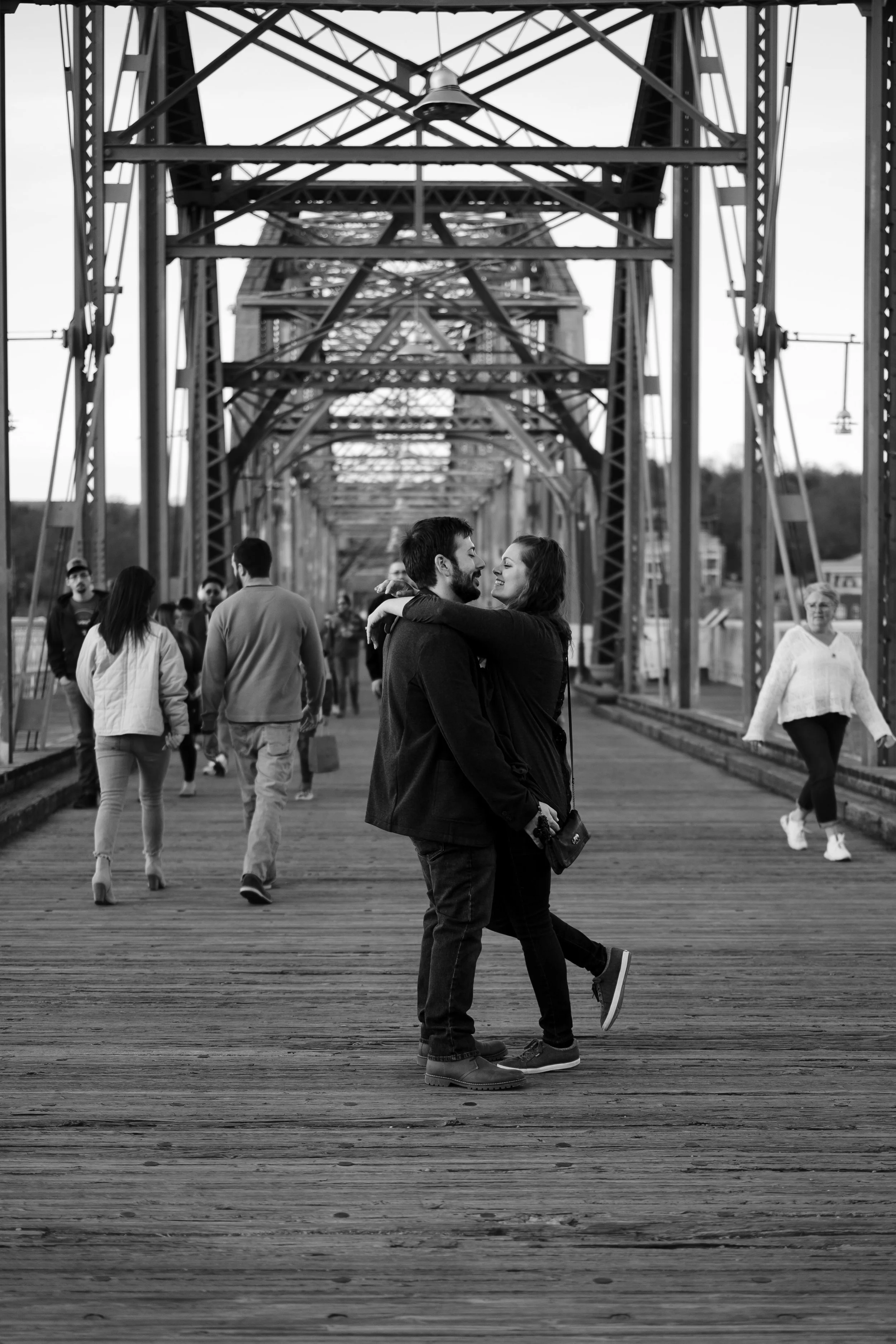 A romantic couple hugging on a bridge with people walking in the background, black and white photo.