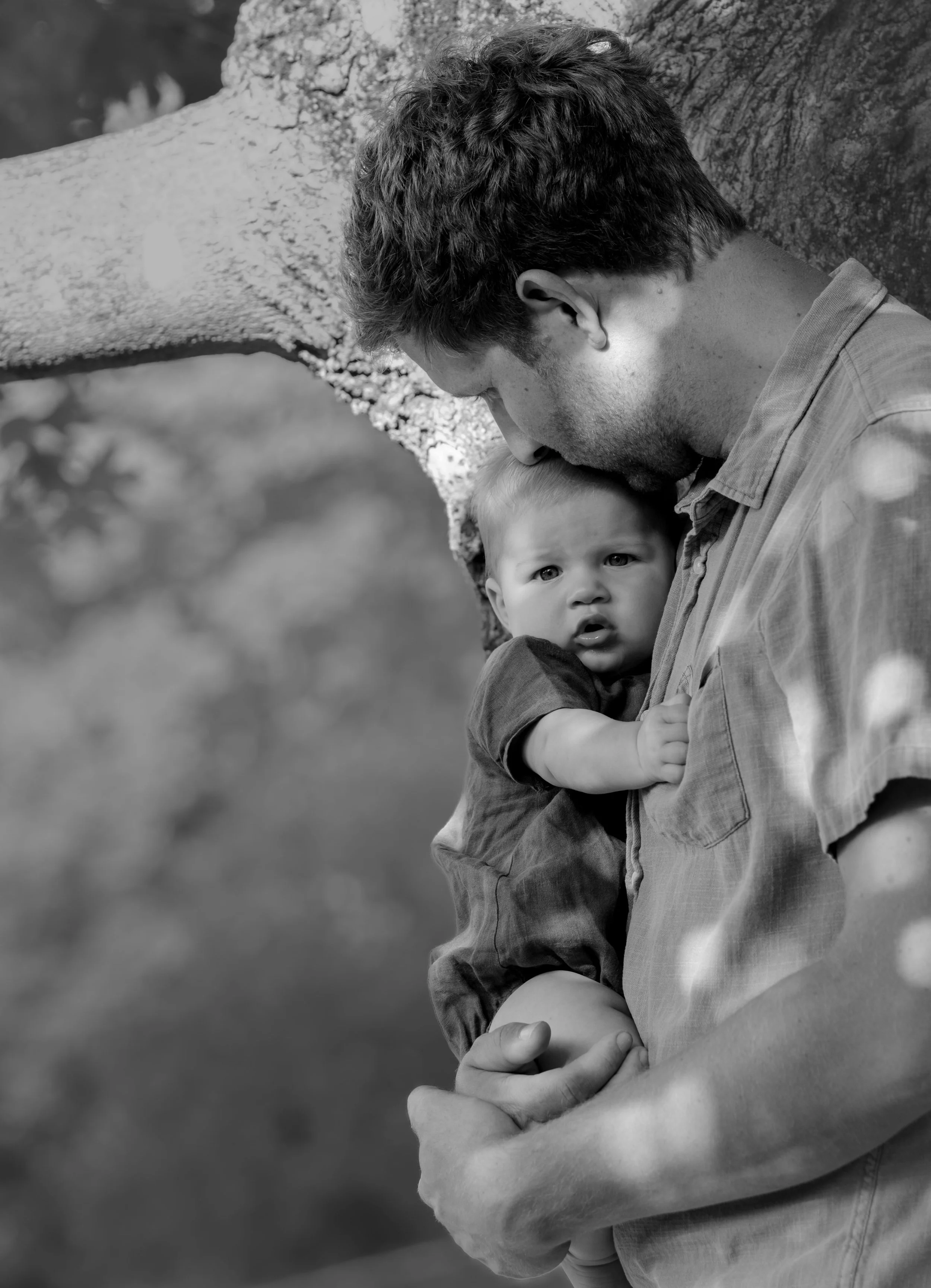 A man holding a young child, who is resting against his chest outdoors near a tree. The man is looking down, and the child has a curious or concerned expression, with a hand gripping the man's shirt.