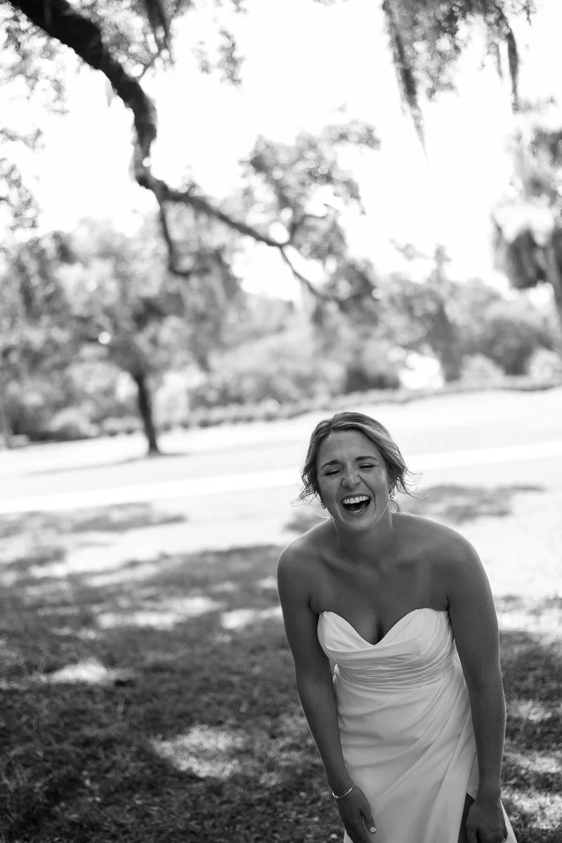 A woman in a strapless dress laughing outdoors under trees in a park on a sunny day, black and white photograph.
