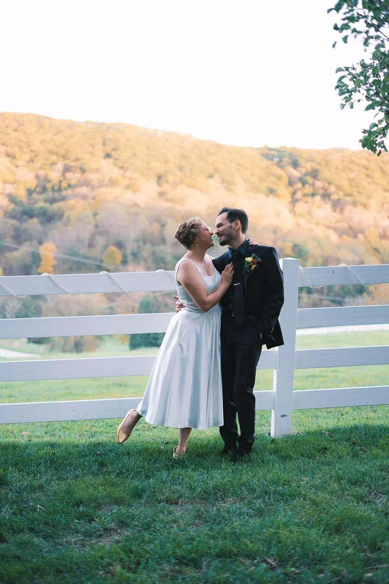 A couple, dressed in wedding attire, shares a romantic moment outdoors next to a white fence with a hill in the background during daytime.