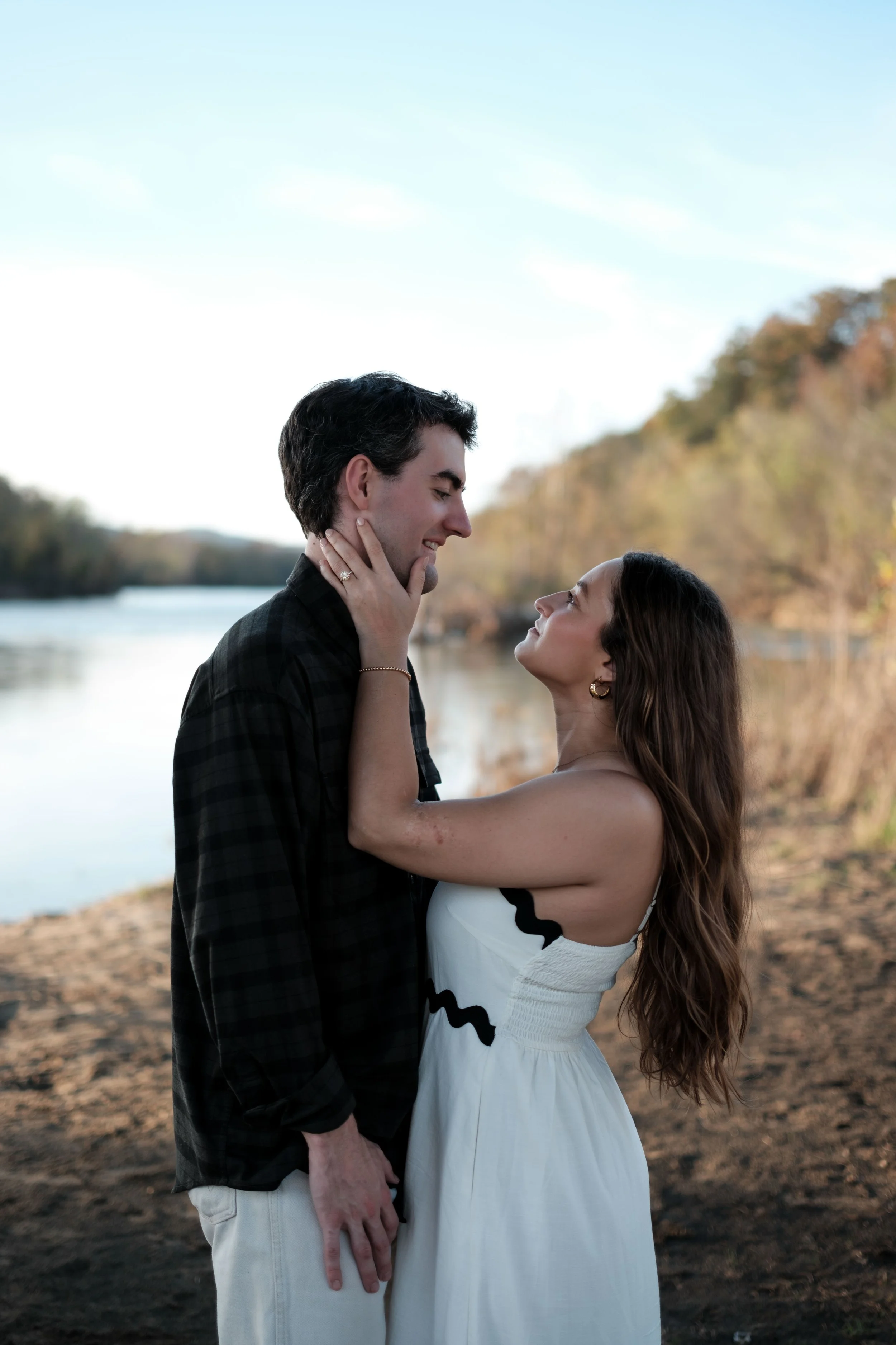 A couple stands outdoors by a river, gazing into each other's eyes, with the woman gently holding the man's face.