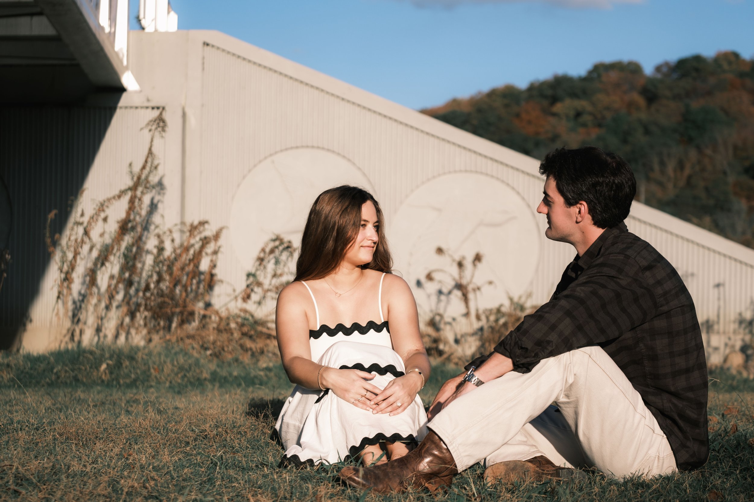 A young man and woman sit on the grass facing each other outdoors. The woman has long brown hair and is wearing a white dress with black trim, while the man has dark hair and is wearing a black plaid shirt and beige pants. They are smiling and lookin