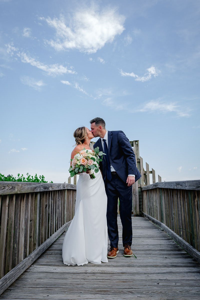 A bride and groom sharing a kiss on a wooden bridge outdoors. The bride holds a bouquet of roses and greenery, and the sky is blue with scattered clouds.