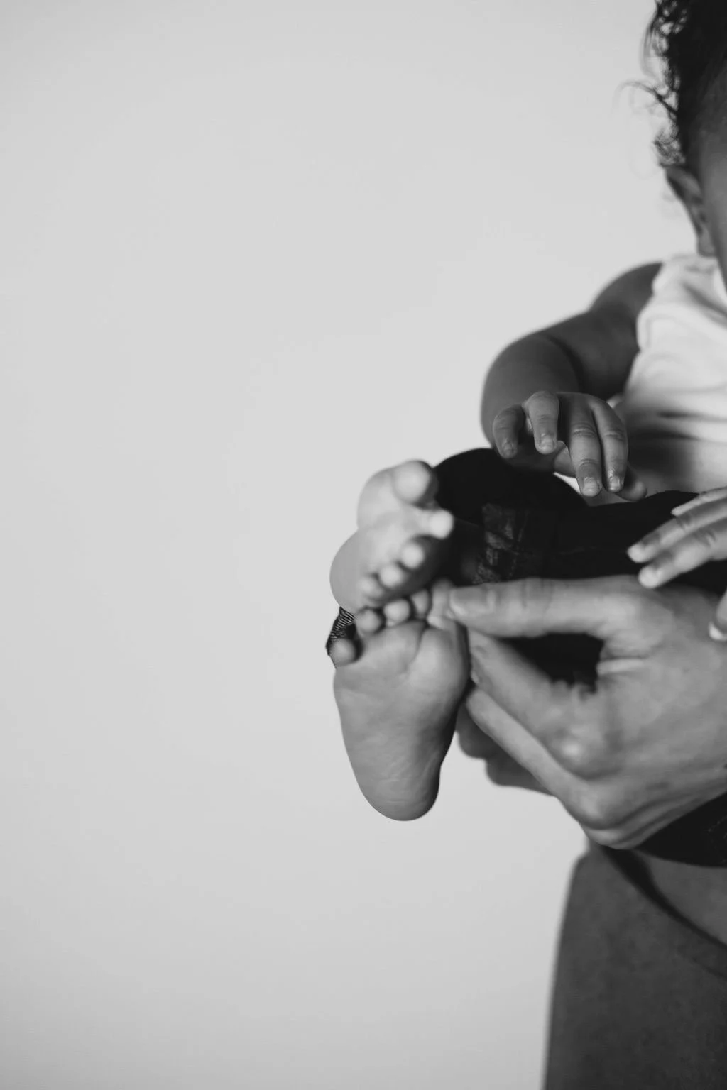 A close-up black and white photo of an adult holding a child's foot, with the child's hand touching it.