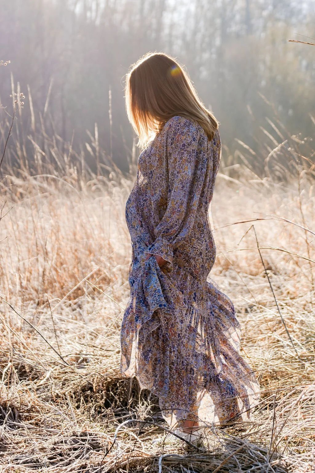 A pregnant woman in a floral dress stands in a field of tall, dried grass during sunset, with her head bowed and hands in her pockets.