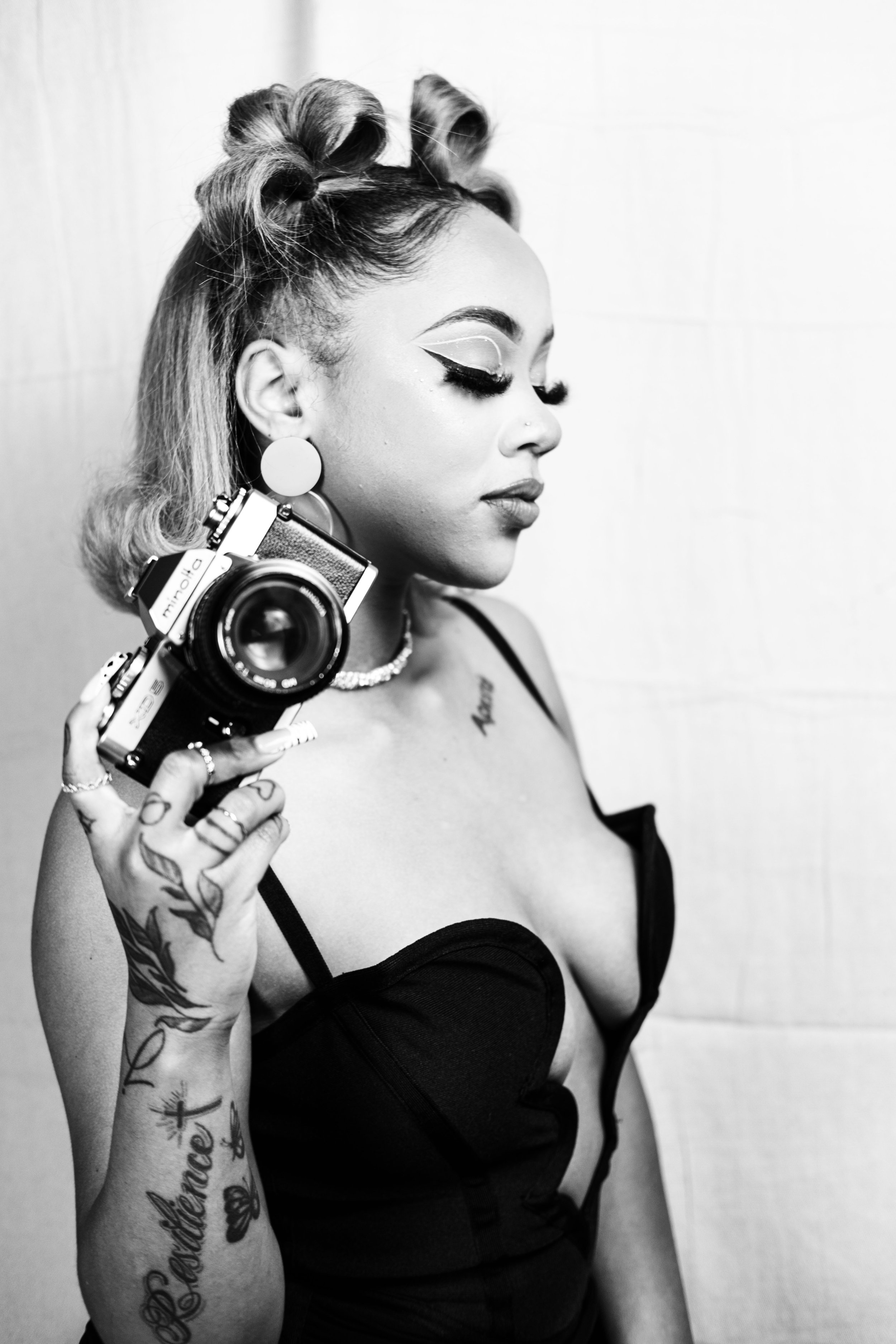 A woman with tattoos, vintage hairstyle, and bold makeup holds a camera to her shoulder, standing against a plain background.