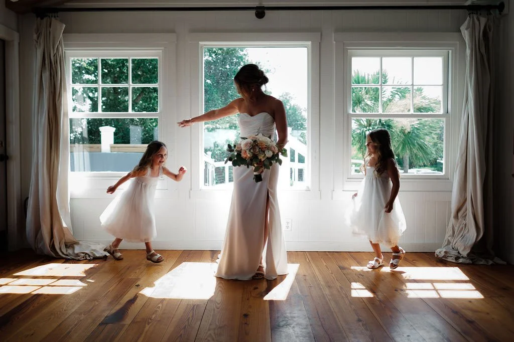 A bride standing in front of three large windows, holding a bouquet, with two young girls in white dresses dancing around her inside a bright room.