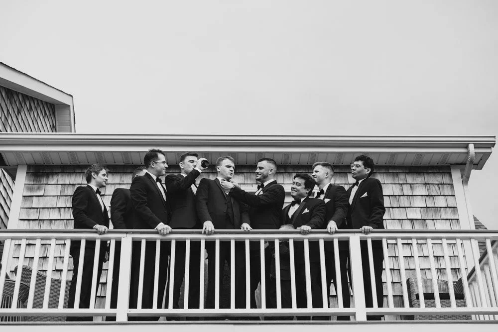 A group of nine men in tuxedos standing on a balcony, some taking photos of each other.