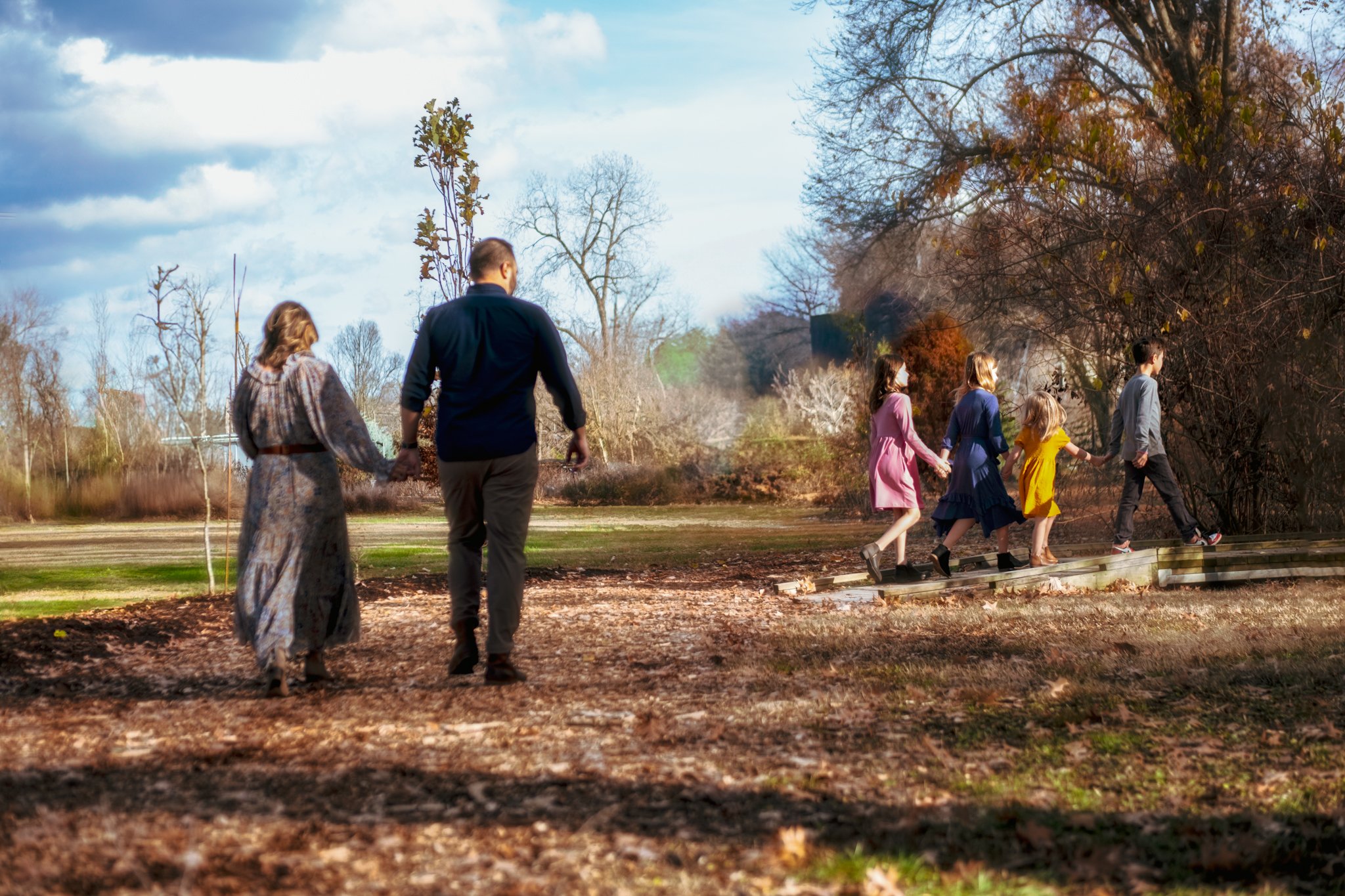 A family of seven walking outdoors in a park during autumn. The adults are holding hands with the children, who are walking on a small wooden bridge surrounded by trees with bare branches.