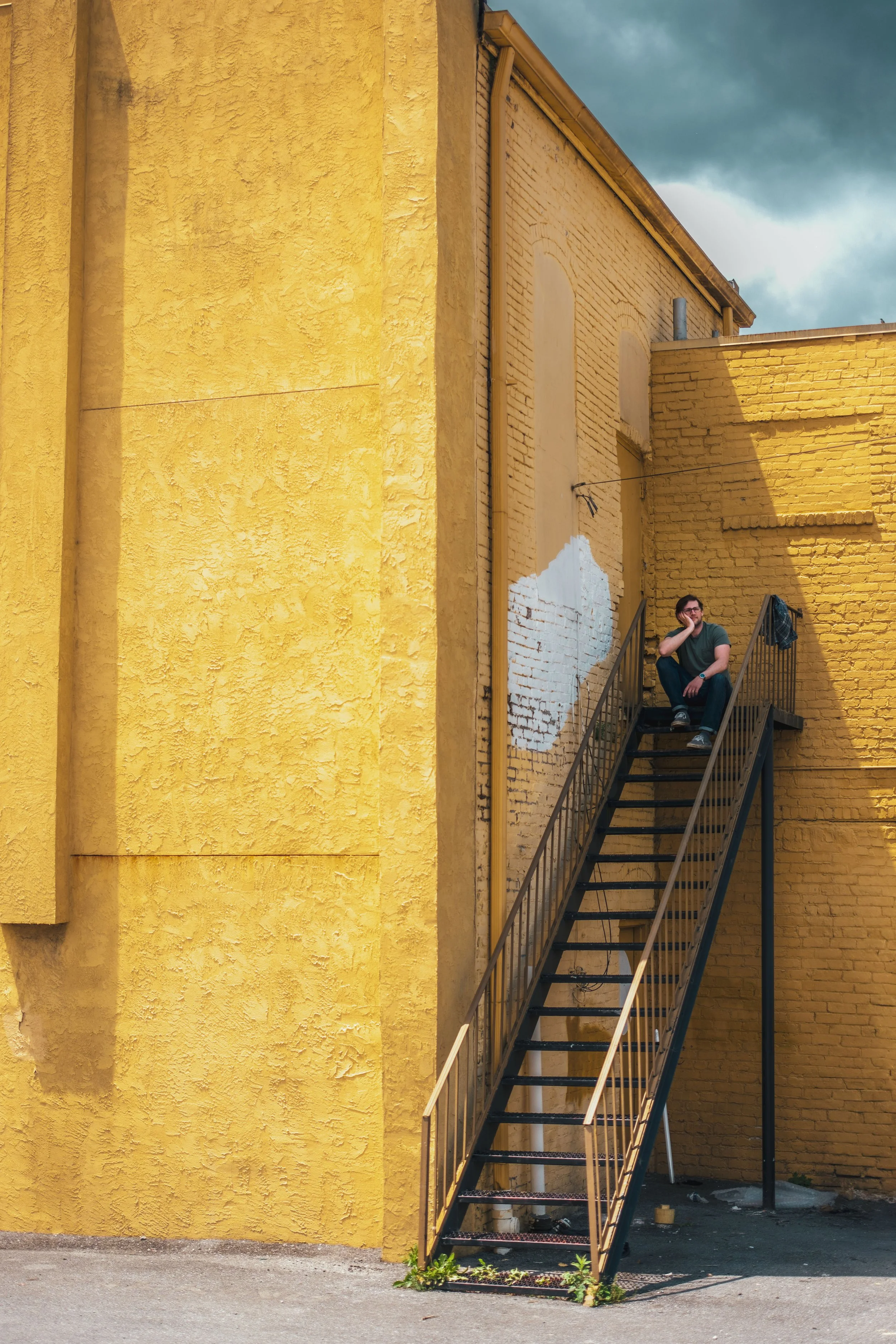 A young man sitting on a metal outdoor staircase attached to a yellow brick building, with a painted white section on the brick wall behind him. He is resting his head on his hand, looking off to the side, under a cloudy sky.
