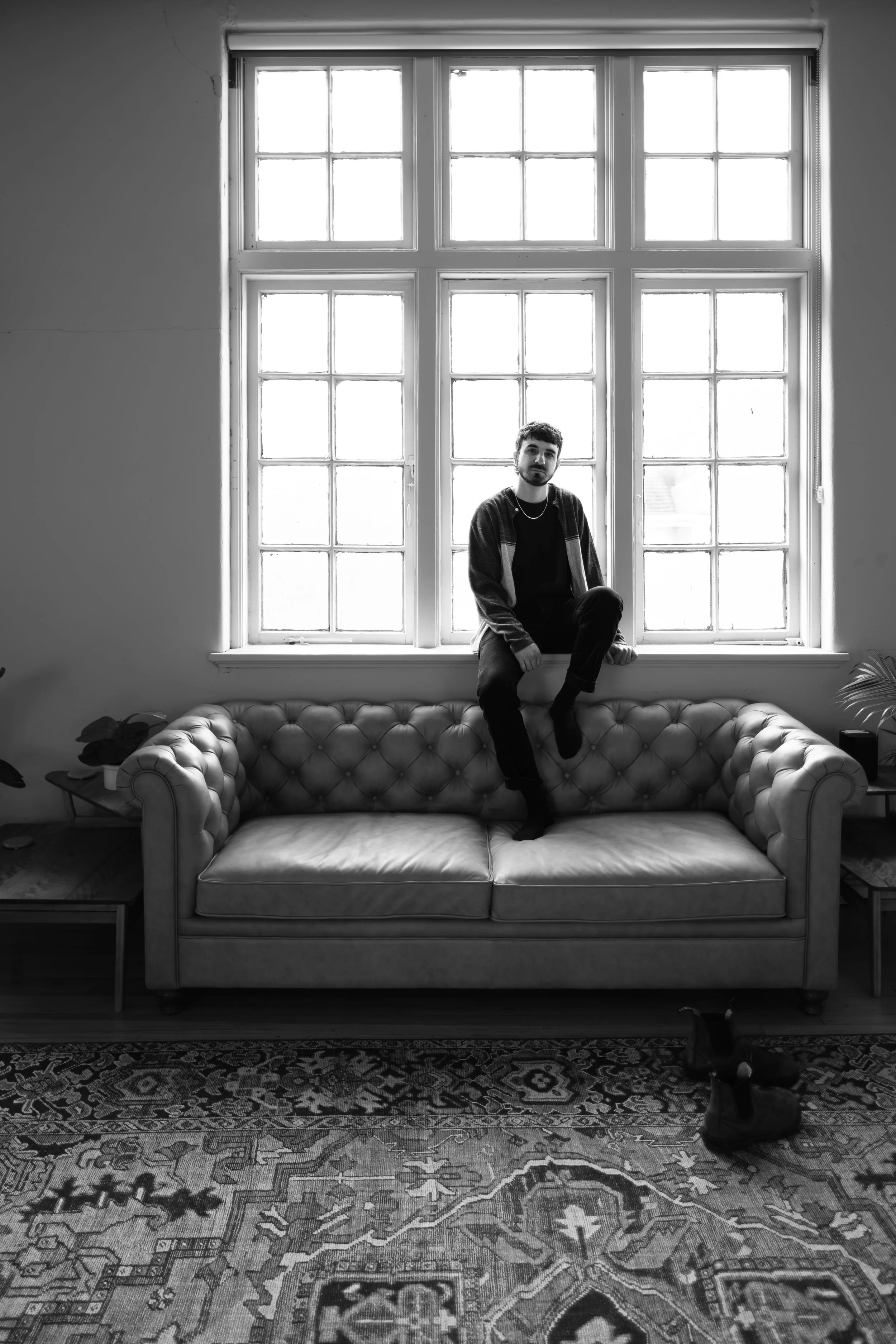 A young man with a beard sitting on a couch in front of a large window, with a pair of boots on the carpet below.