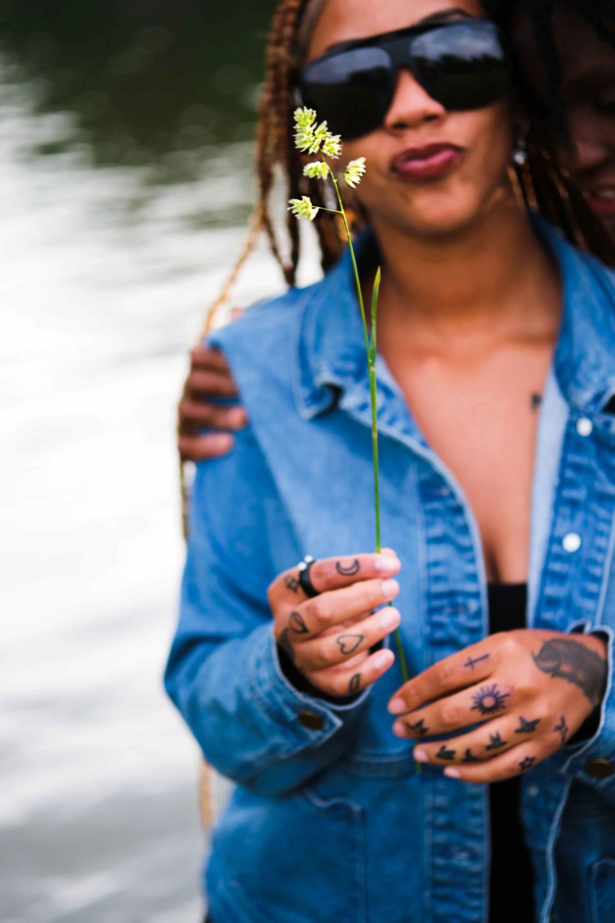 Person in sunglasses holding a small flower, wearing a denim jacket, standing near a body of water.