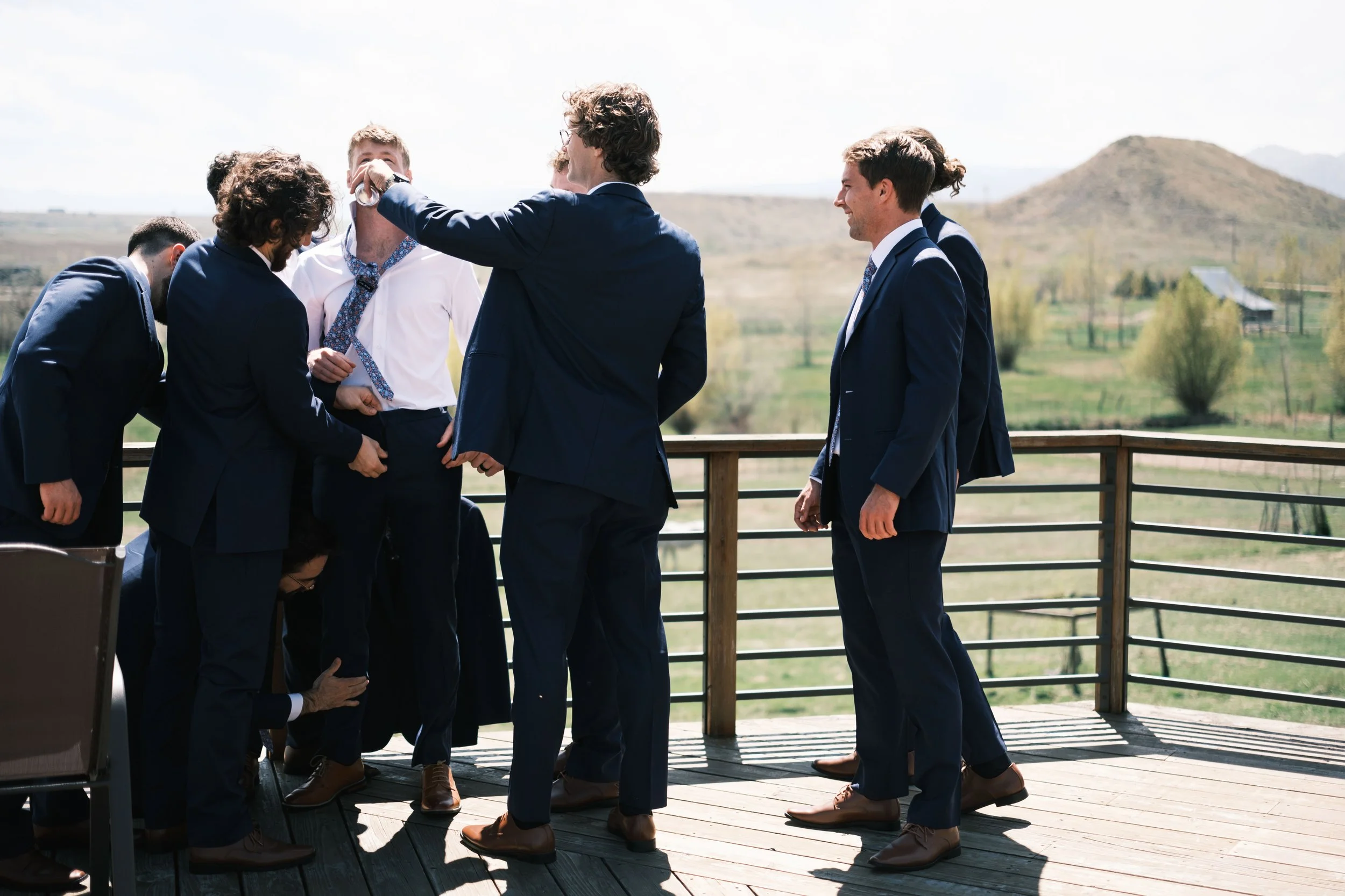 Group of men in suits on a wooden outdoor deck, some smiling and one adjusting another's tie, with a scenic rural landscape in the background.