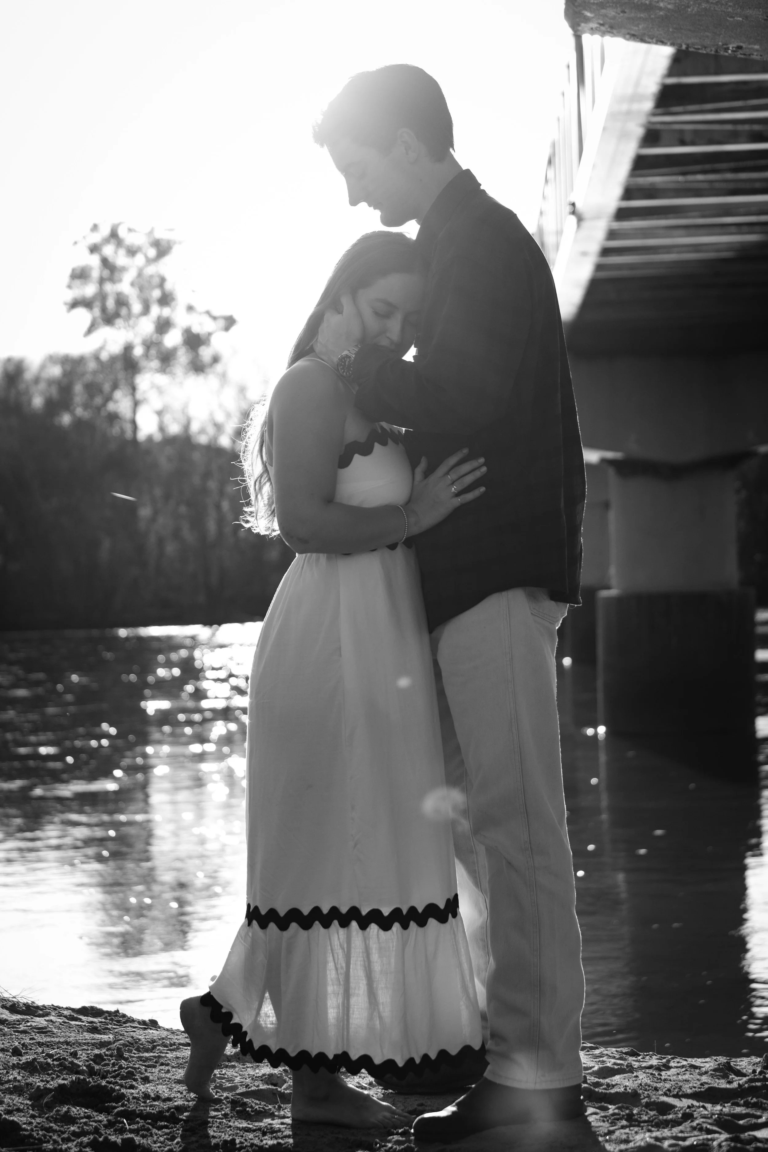 A black and white photo of a couple embracing on a beach under a bridge with water nearby, backlit by the sun.