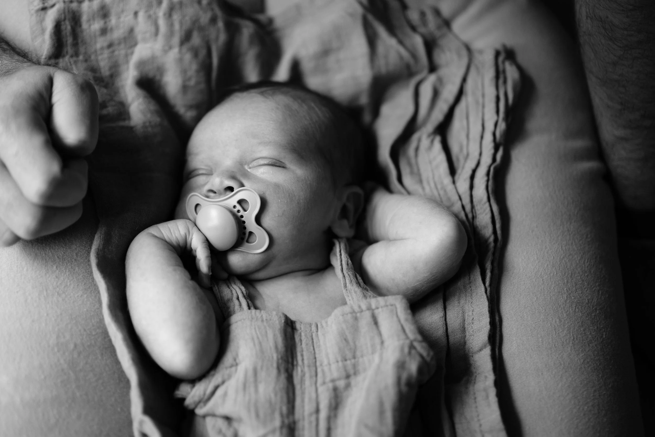 A sleeping baby with a pacifier in their mouth, lying on a person's lap, with their hands behind their head, in black and white.
