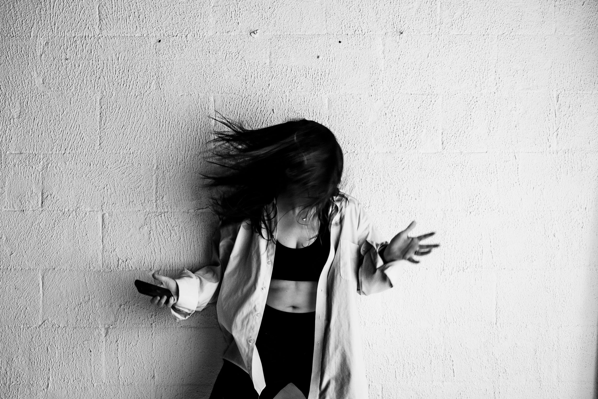 Black and white photo of a woman dancing or moving to music while holding a phone, with hair swinging, against a textured wall.