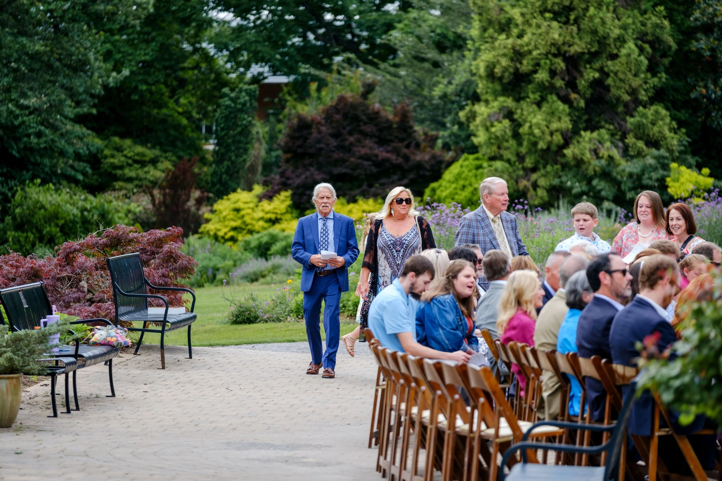 People attending an outdoor event in a garden with green trees and colorful bushes, some seated at wooden chairs and others standing, dressed in semi-formal attire.