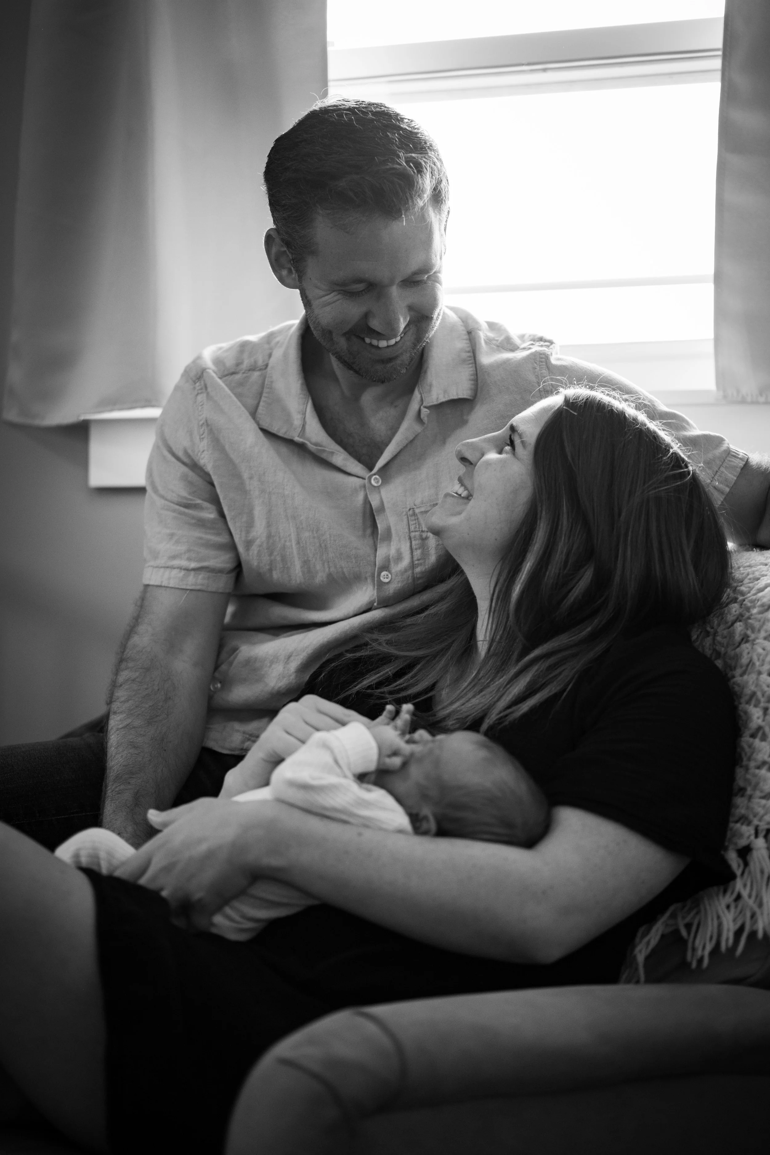A joyful family moment in a hospital, with a woman holding a newborn baby, a man leaning over her smiling, in a softly lit room.