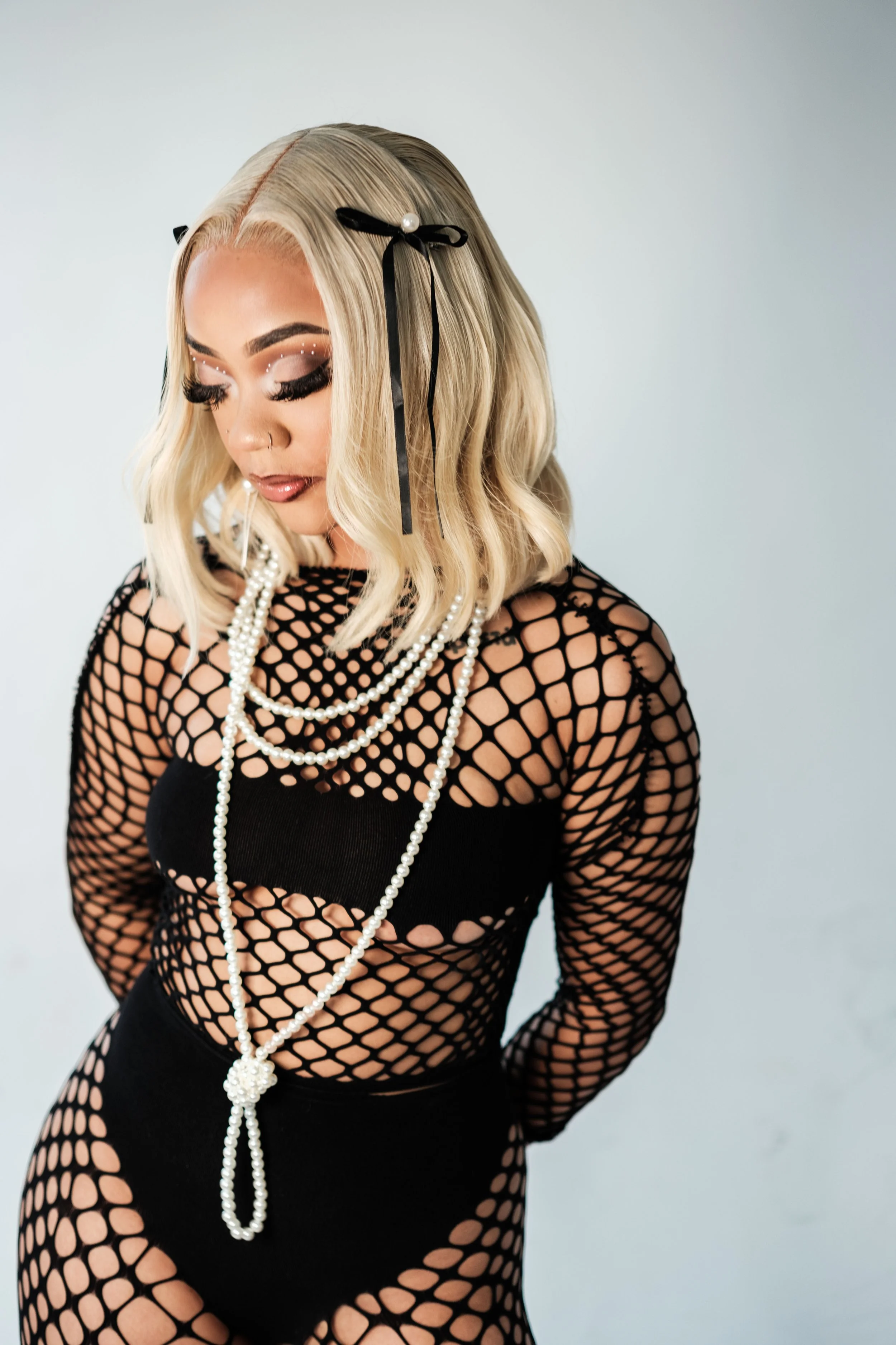 Woman with blonde hair in waves, wearing black fishnet outfit and layered pearl necklaces, with hair accessories and makeup with dramatic eyelashes and rhinestones, posed against a neutral background.