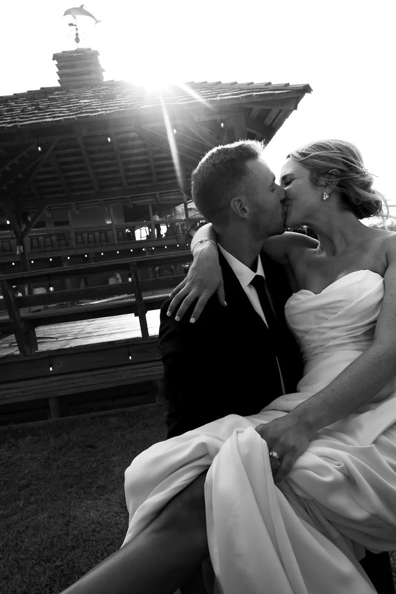 Black and white photo of a newlywed couple kissing outdoors, with a dock and building in the background during sunset.