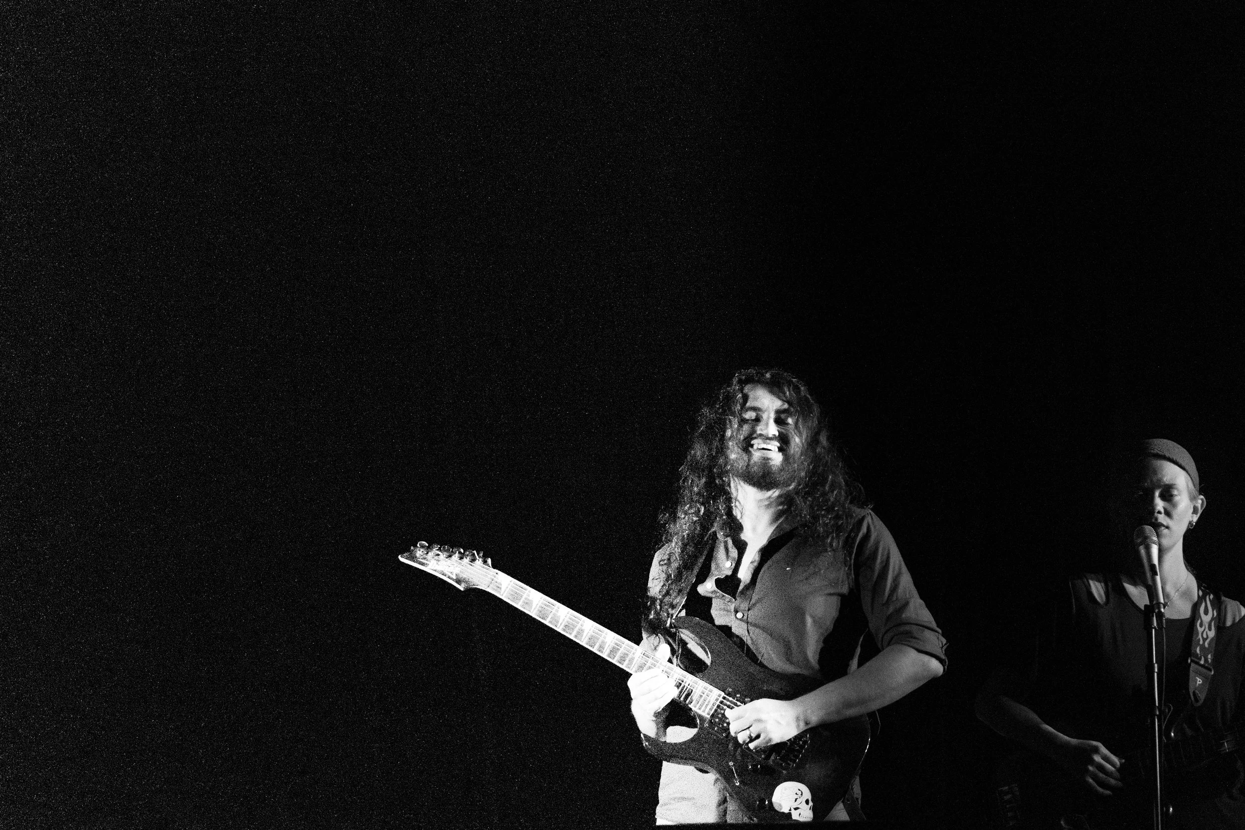 A man with long curly hair playing an electric guitar; a woman next to him singing into a microphone, both on stage during a performance.