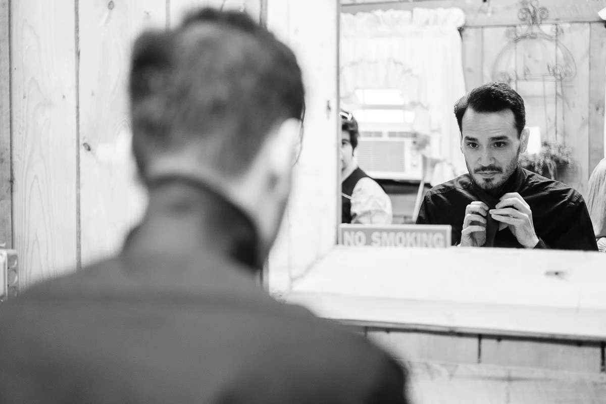 A man adjusting his collar in front of a mirror, with another man’s reflection visible in the mirror. The setting appears to be a barber shop or salon, with a 'No Smoking' sign on the mirror and an air conditioning unit in the background.