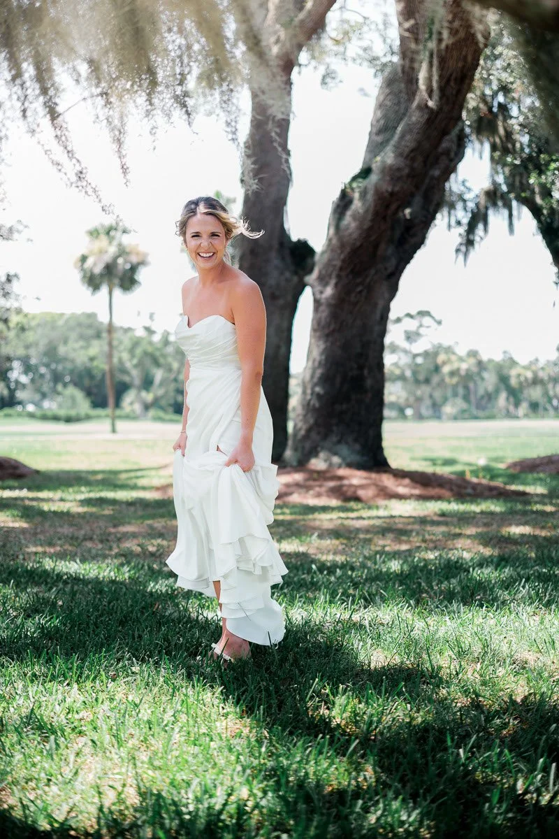 A woman in a white dress smiling and walking barefoot in a park with large trees and green grass.