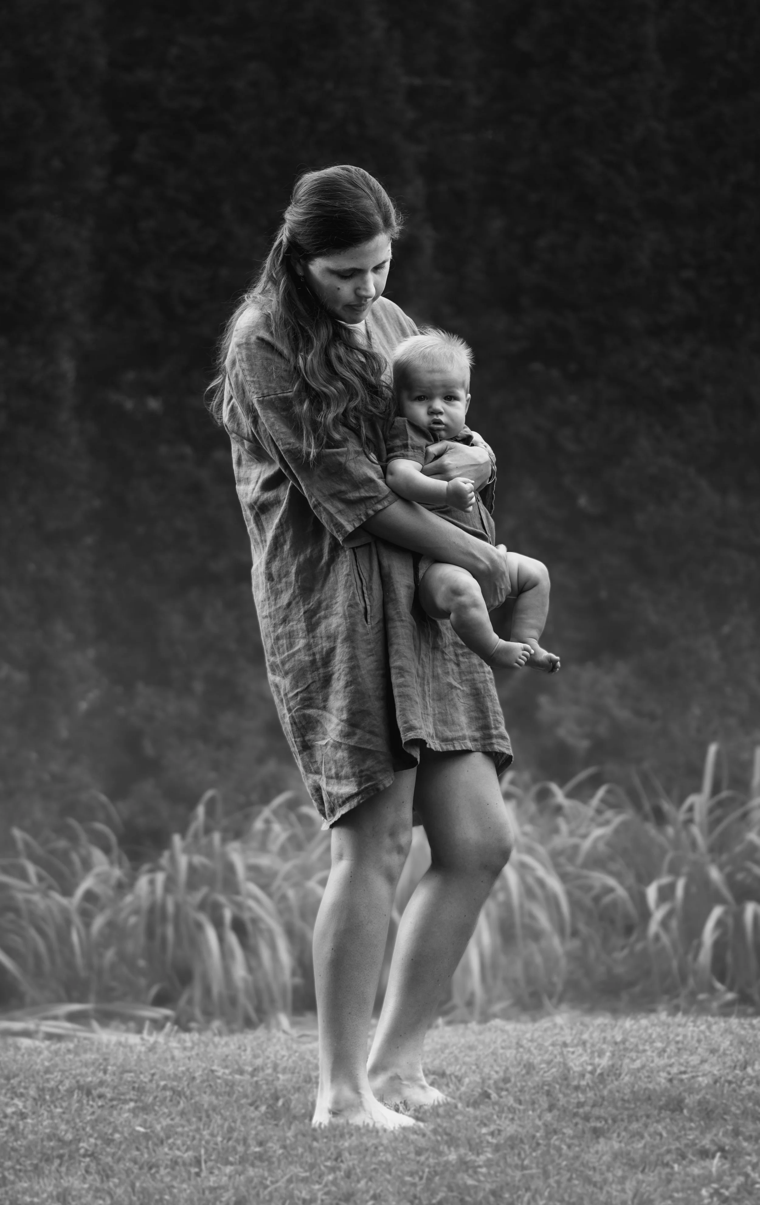 A woman in a dress holding a young child outdoors in a grassy area with flowers and trees in the background, in black and white.