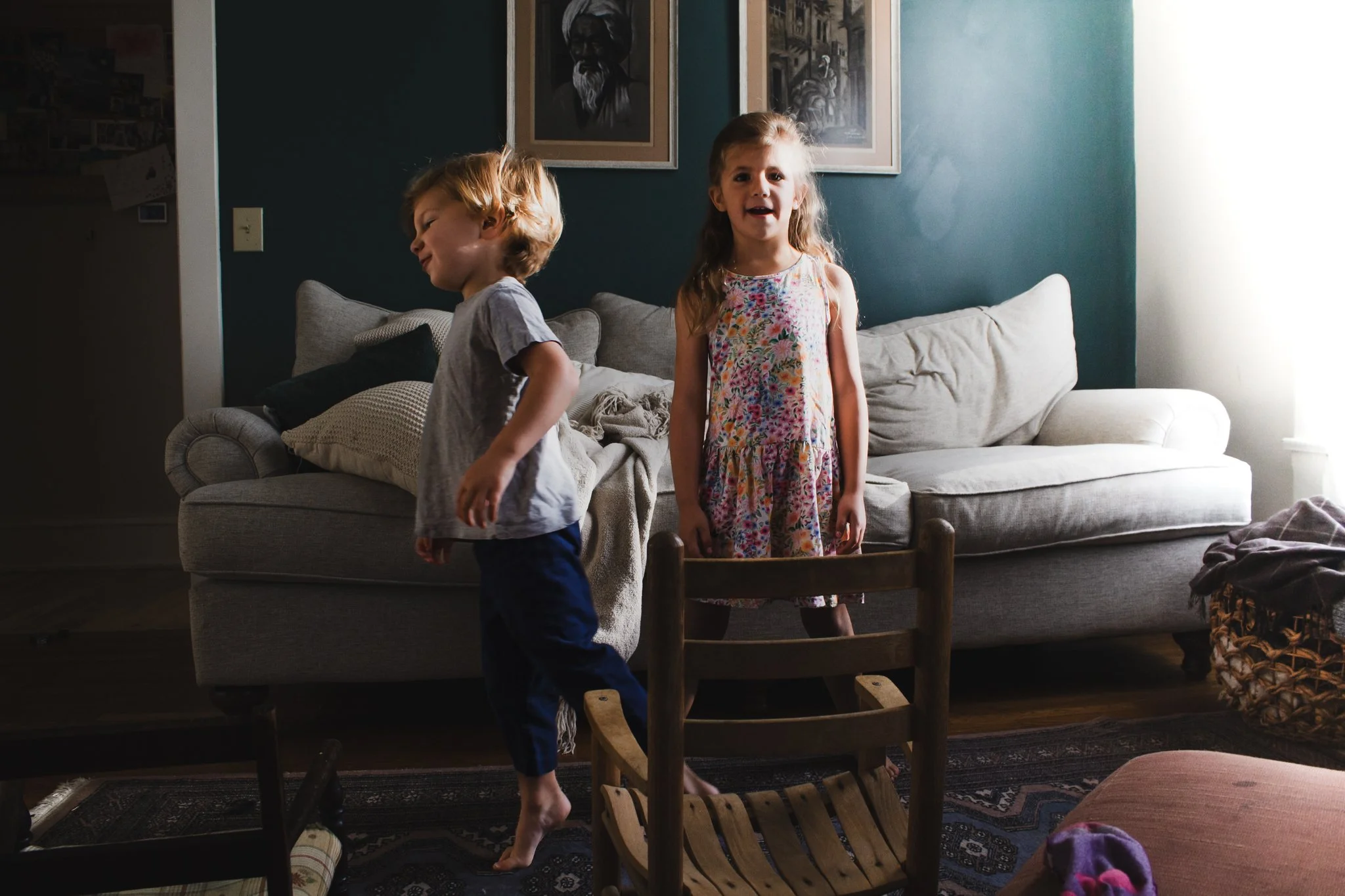Two young children, a boy and a girl, playing in a living room. The girl is standing on a small wooden chair, looking at the camera with a smile. The boy is walking past her, facing away. There is a light-colored sofa with pillows and a blanket behin