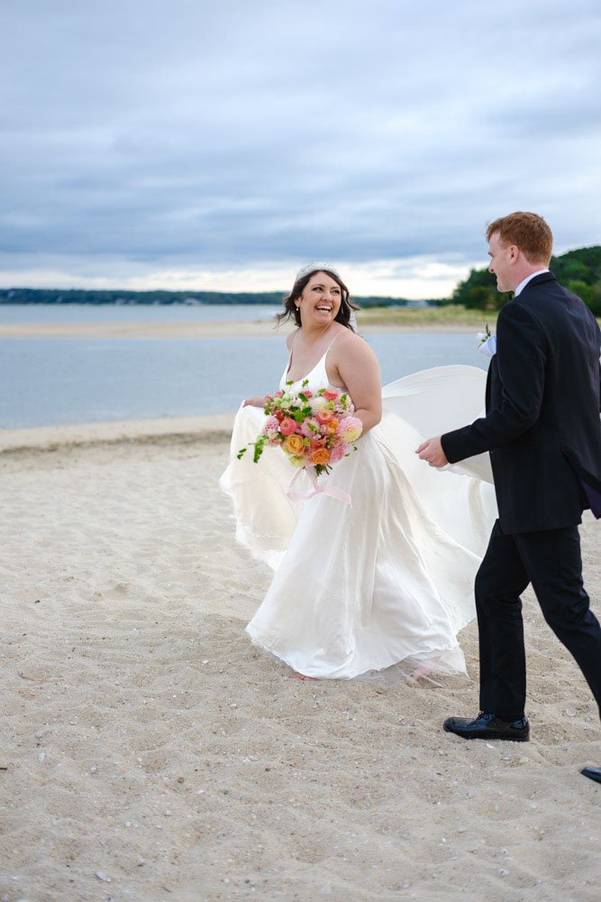 A bride in a white wedding dress holding a colorful bouquet, smiling as she walks on the beach with a groom in a black suit, near a body of water under a cloudy sky.