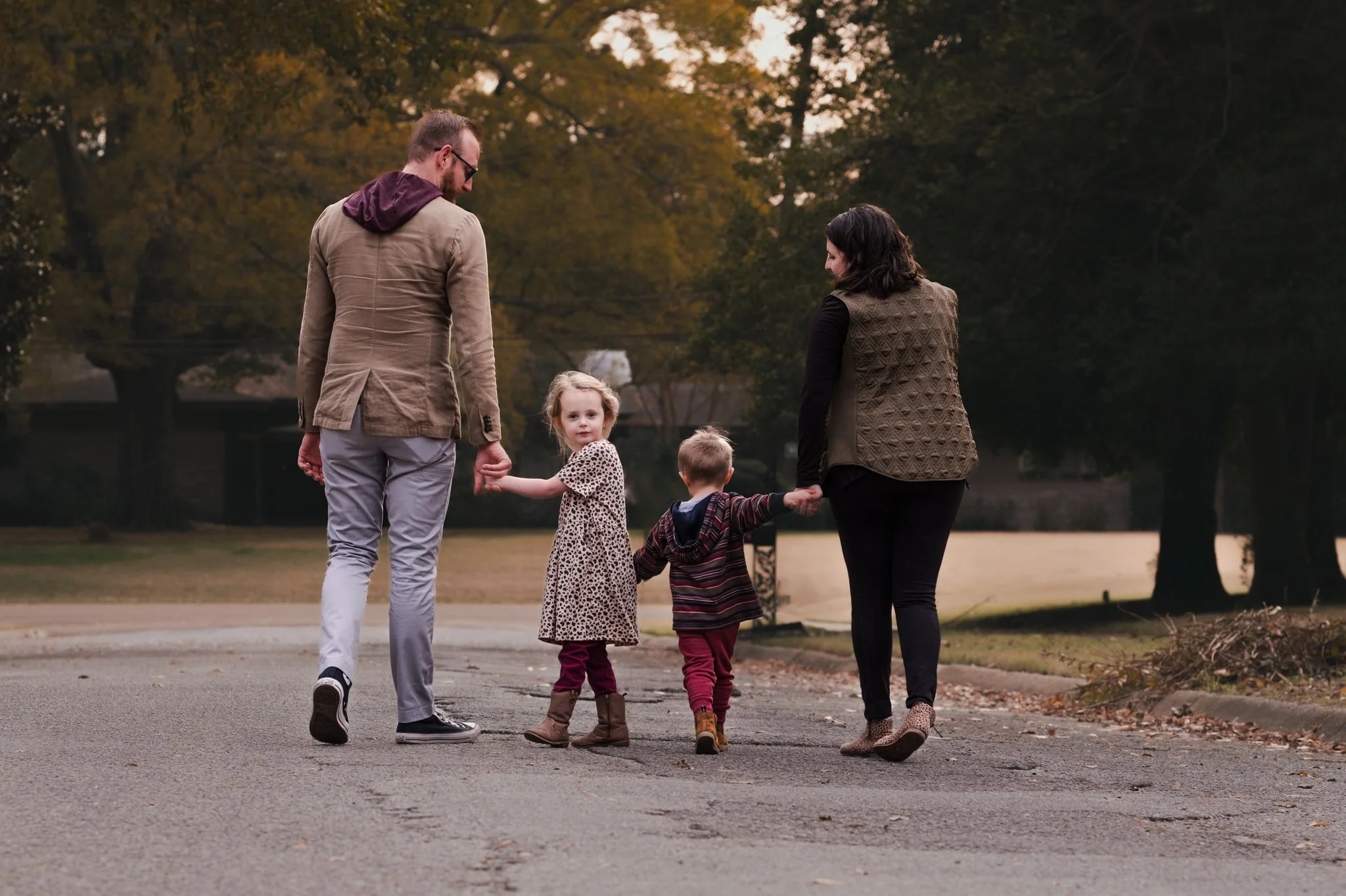 A family of four walking on a paved path in a park during autumn, holding hands and looking back at the camera. The family includes two adults and two children, a girl and a boy. The trees in the background have fall foliage, and the scene appears to