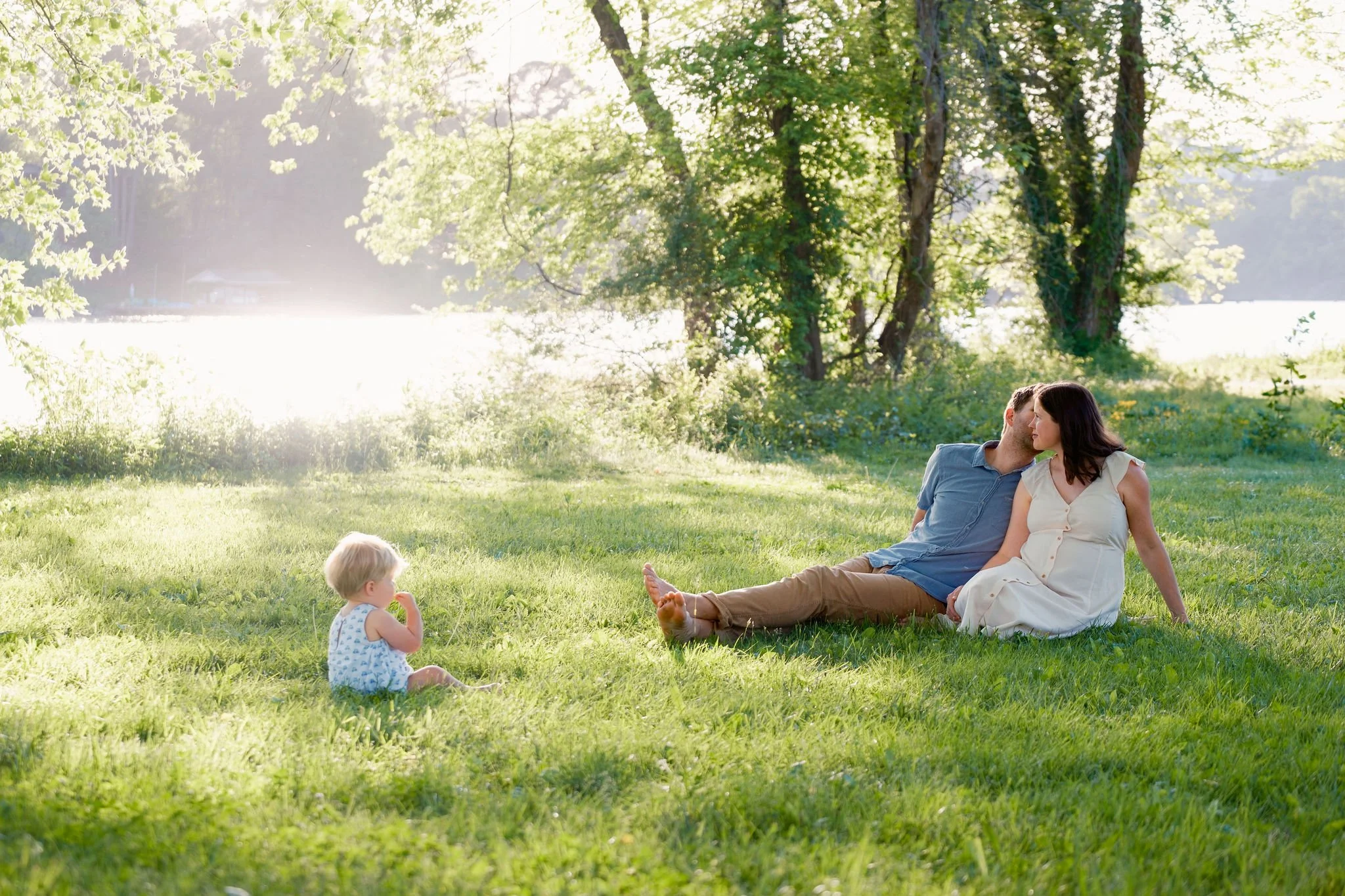 A couple sitting on the grass near a lake, leaning close to each other, with a small child sitting nearby in a park-like setting with trees and sunlight filtering through.